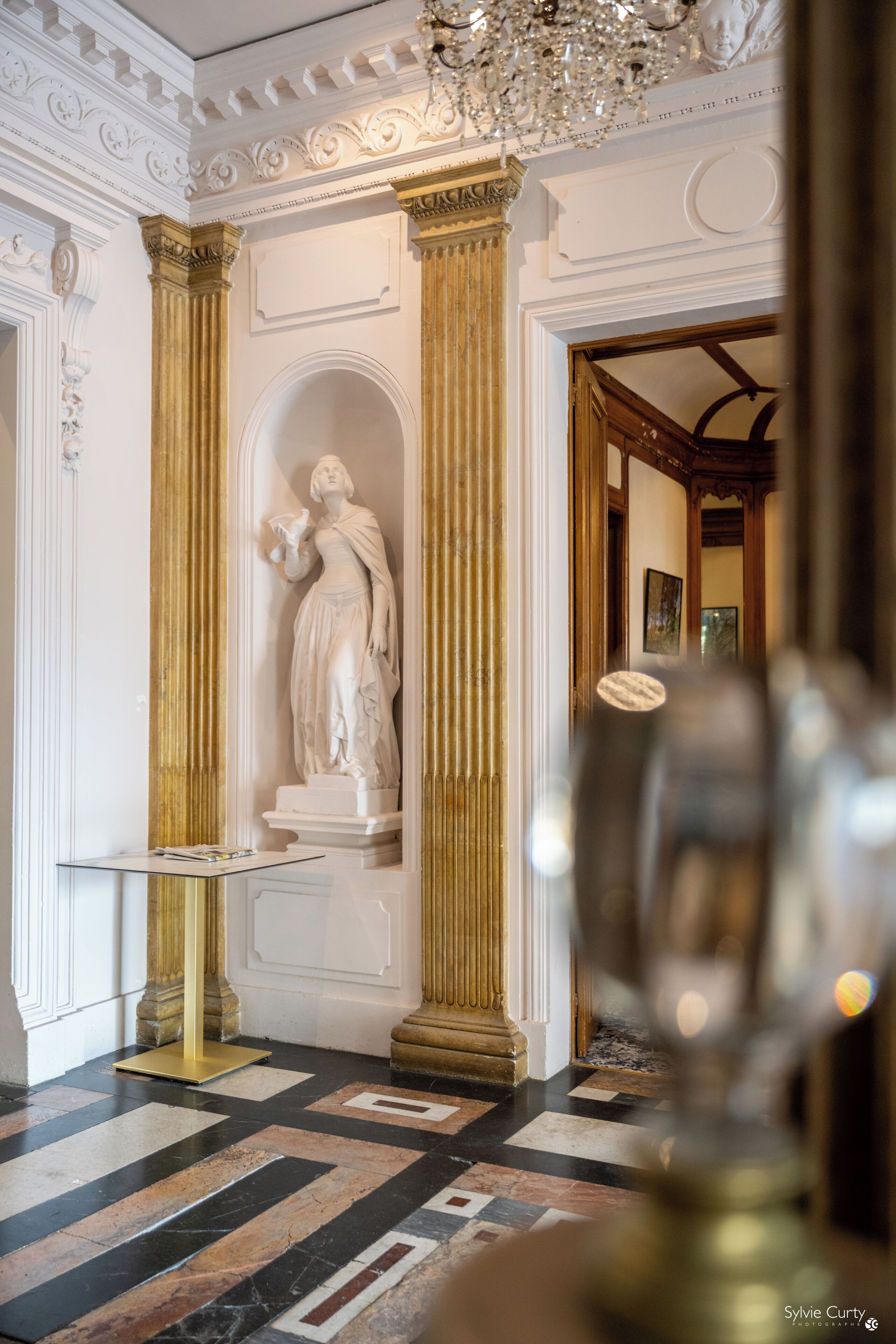 A grand hallway with a marble floor, ornate columns, and a statue.
