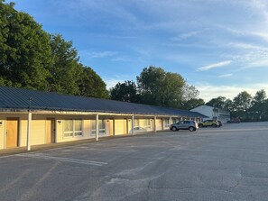 A motel with a row of rooms, a parking lot, and a large building in the background. - Moonlight Inn (Brunswick)