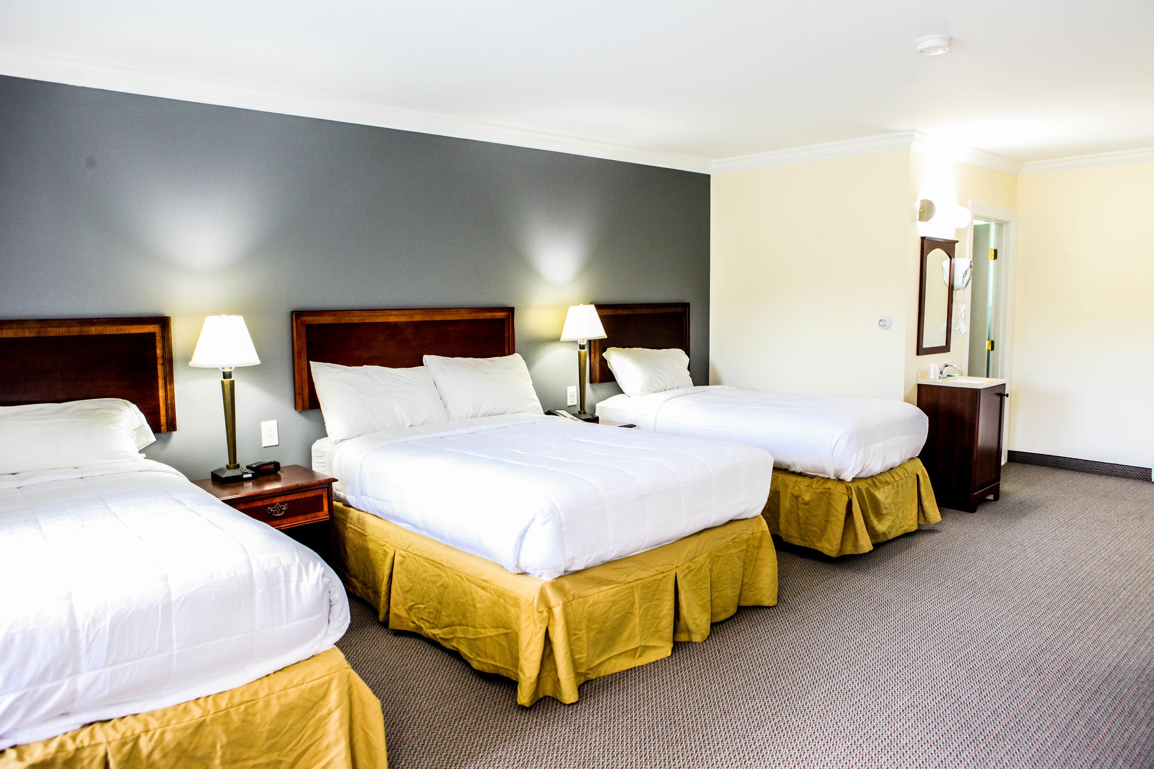 Ein Hotelzimmer mit drei Betten, jeweils mit einem Holz-Kopfteil, weißen Laken und einem gelben Bettschürze. An den Betten stehen Nachttische mit Lampen, ein Spiegel und ein kleiner Holzkasten.