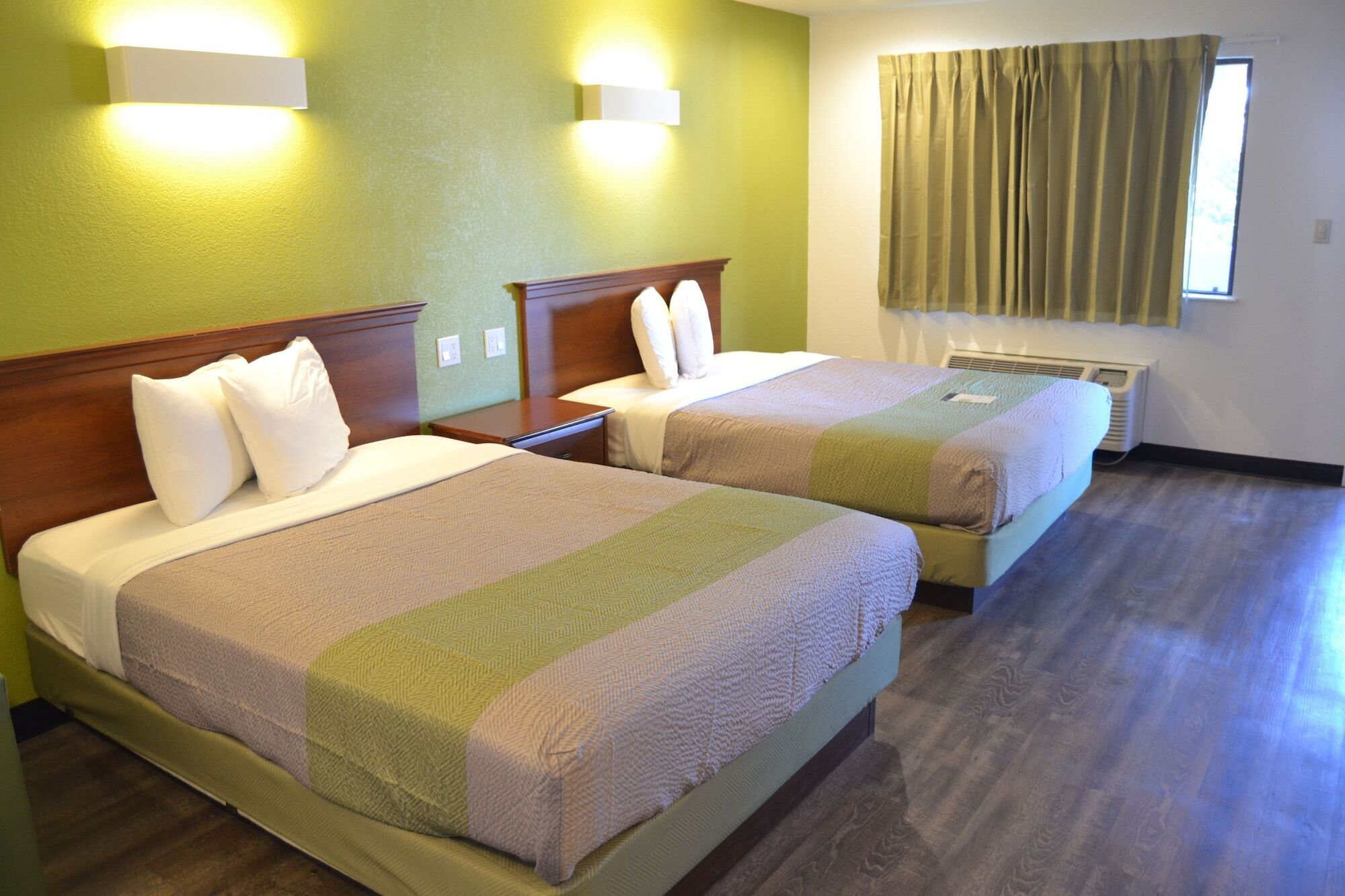 A hotel room with two beds, each with a wooden headboard, white bedding, and a green and white striped blanket. There are wall-mounted lights and a window with curtains.