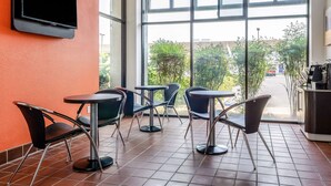 A café with a TV mounted on the wall, several tables and chairs, and a view of the outdoors through large windows.