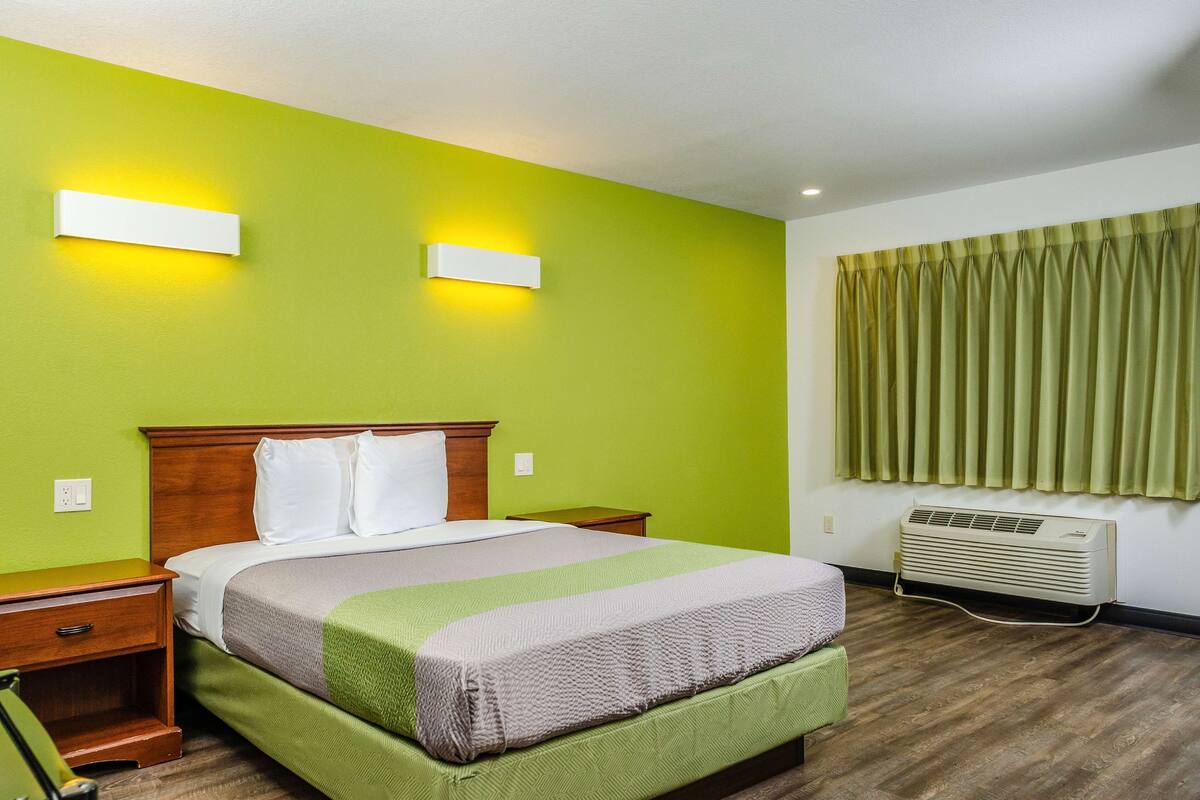 A hotel room with a green wall, a wooden bed frame, a green and white bedspread, two bedside tables, a green curtain, and a wall-mounted air conditioner.