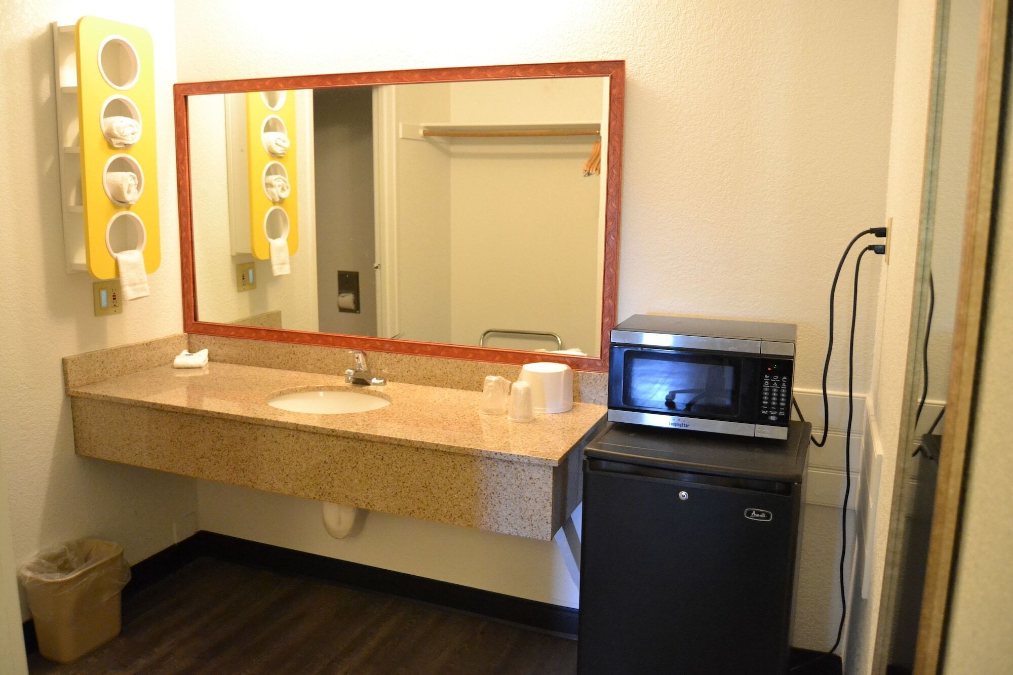 A bathroom with a granite countertop sink, a mirror, a towel dispenser, a microwave, and a trash bin.