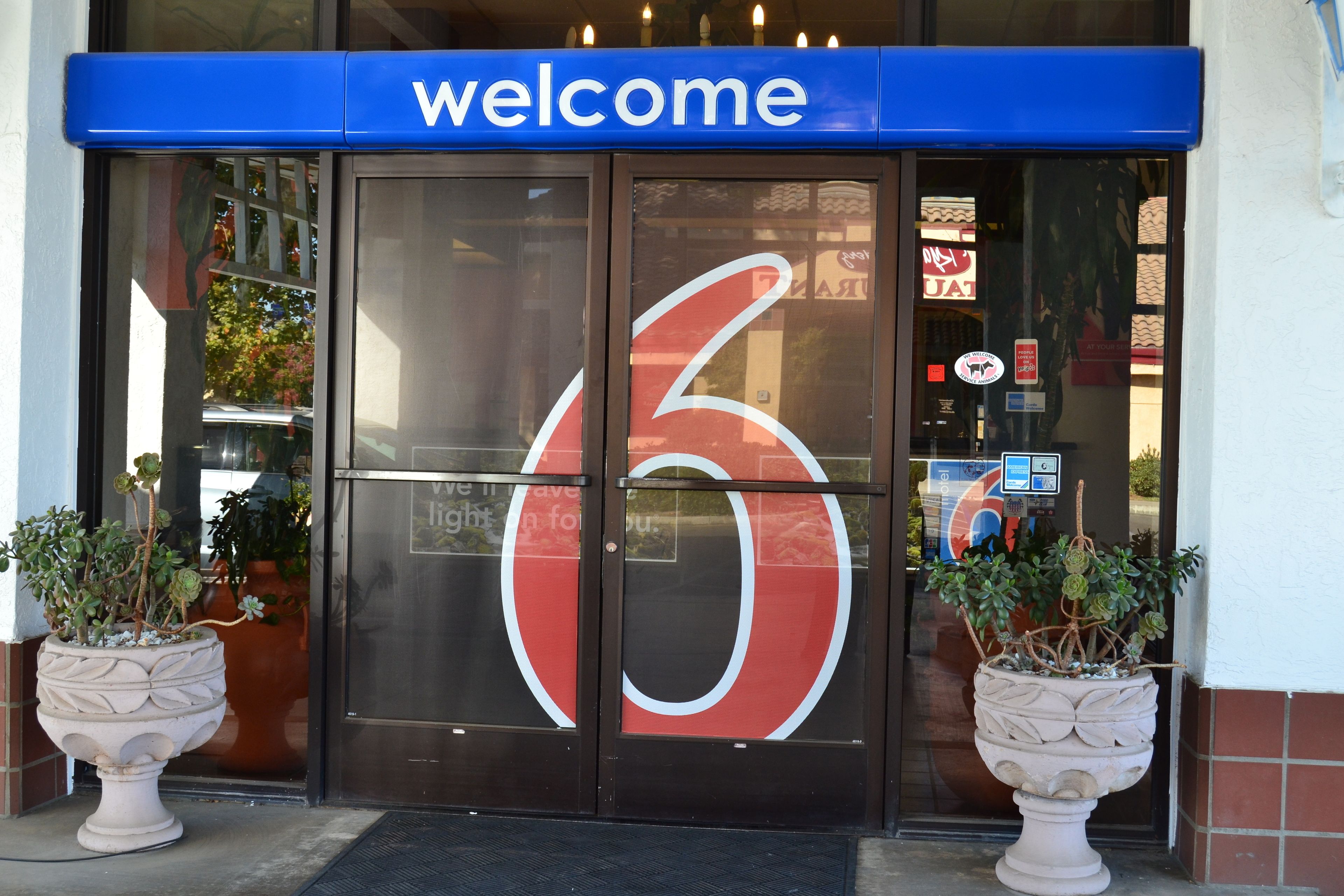 A hotel entrance with a large '6' logo, a 'welcome' sign, and two potted plants.