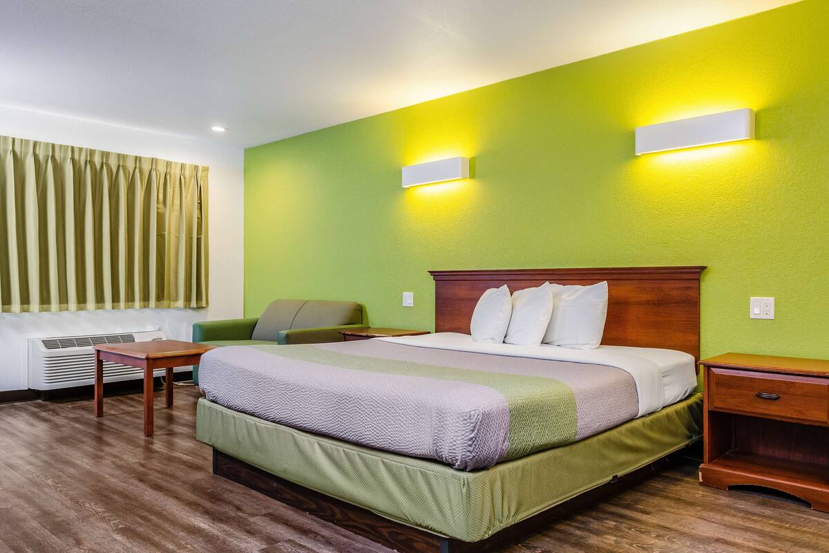 A hotel room with a large bed, a green wall, two bedside lamps, a wooden nightstand, a green armchair, and a window with curtains.