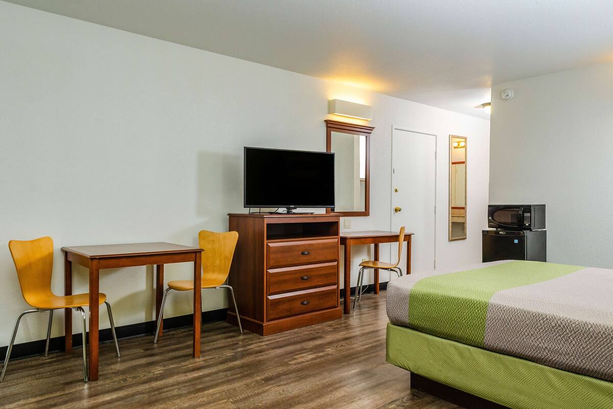 A hotel room with a wooden desk, two chairs, a television, a dresser, a bed with a green and beige bedspread, and a microwave.