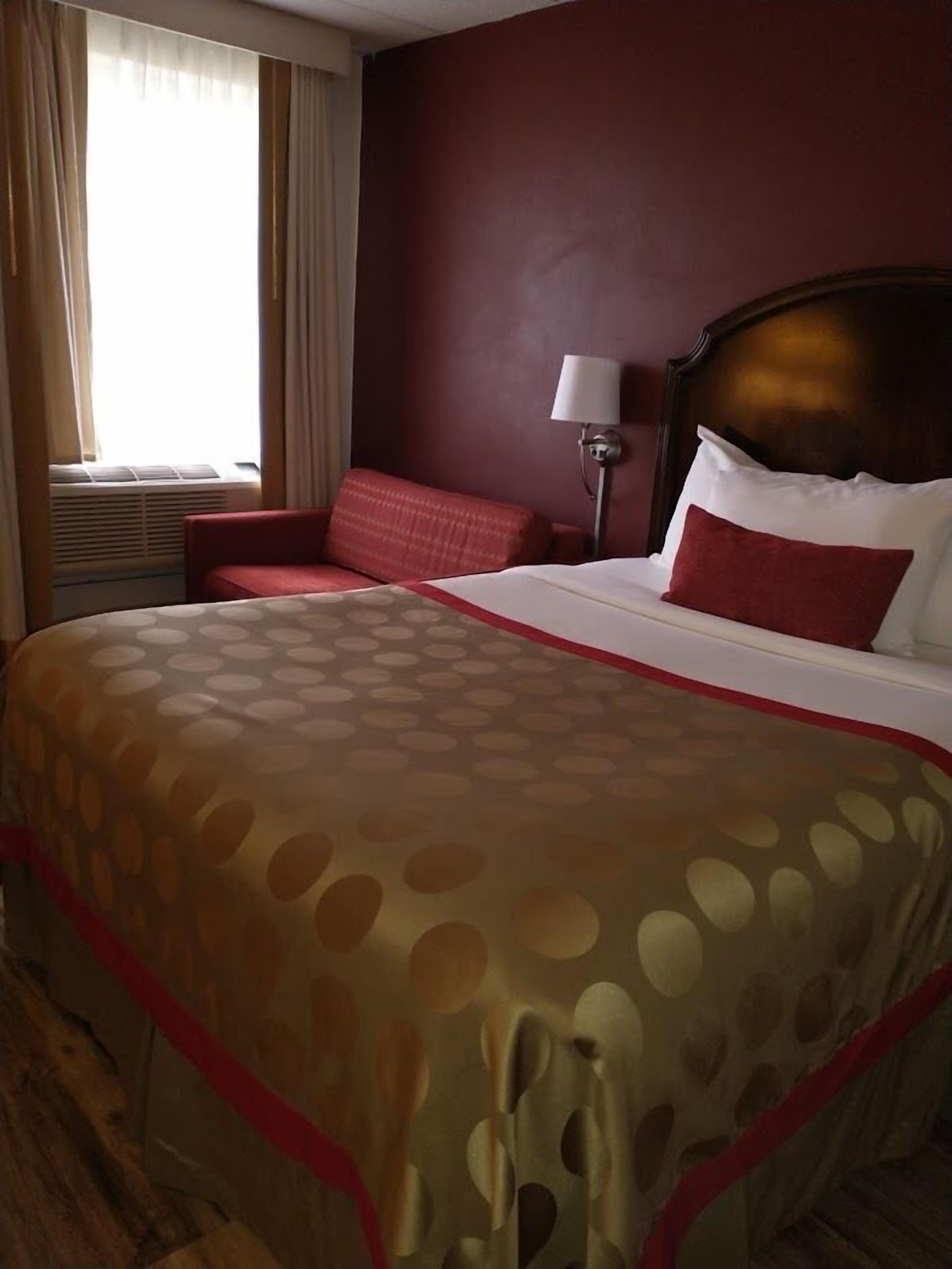 Una camera d'albergo con un grande letto, una poltrona rossa, un comodino con una lampada e un condizionatore d'aria.