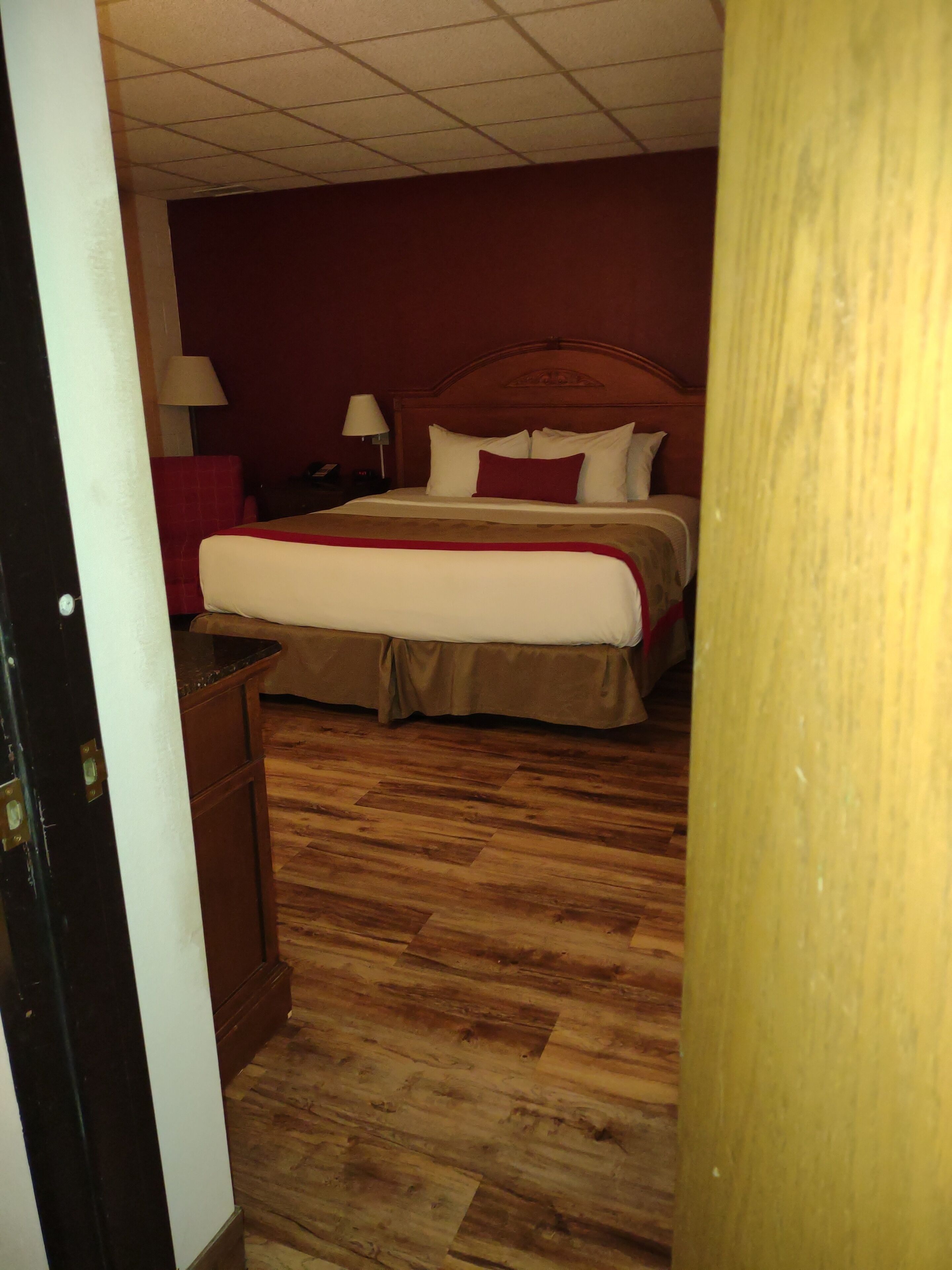 A hotel room with a wooden floor, a bed with white and red bedding, a nightstand with a lamp, and a red chair.