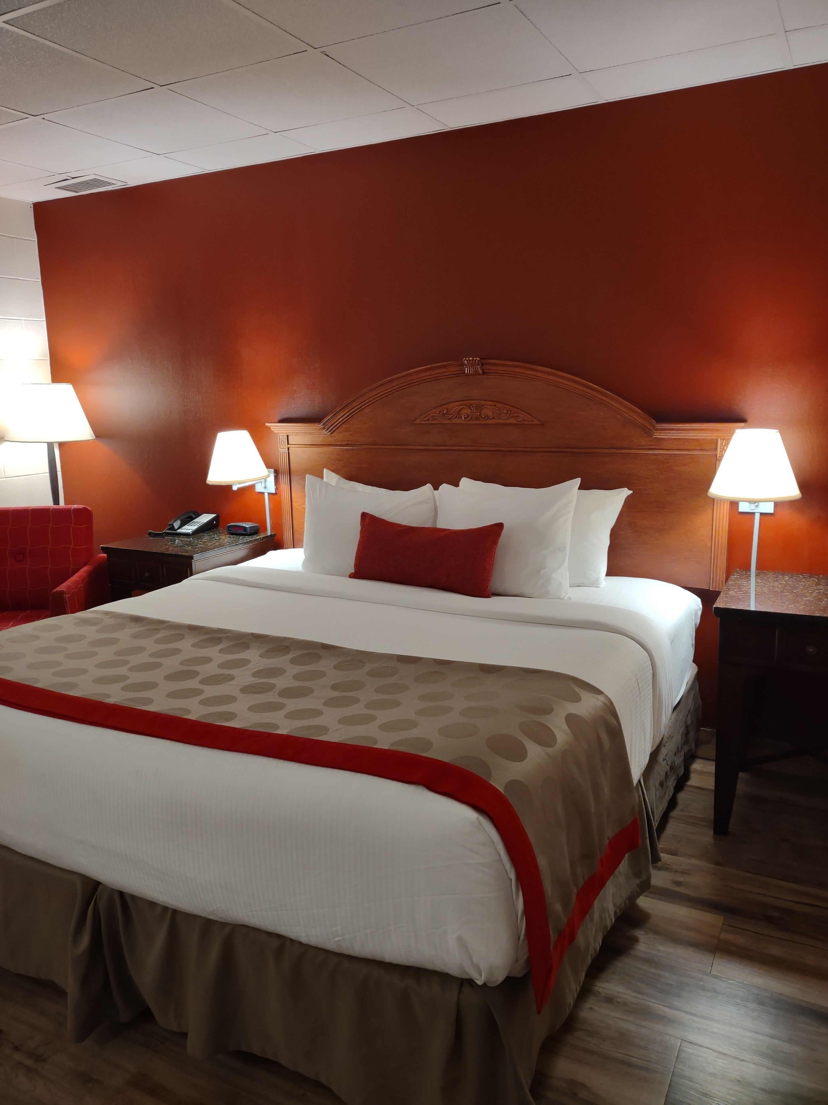 A hotel room with a large bed, two bedside tables with lamps, a red chair, and a wooden floor.
