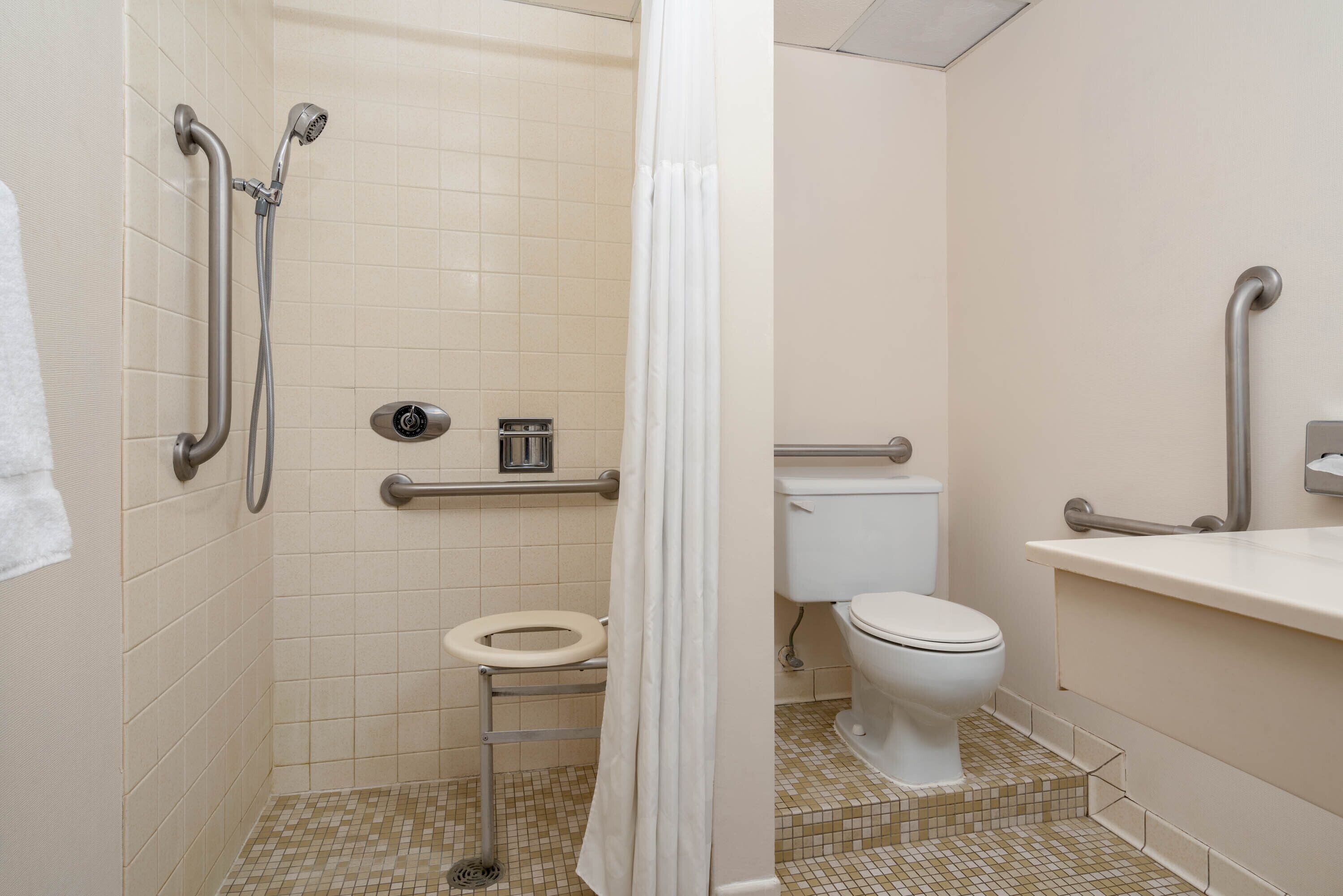 Un baño pequeño y accesible con inodoro, barras de apoyo, ducha con cabezal de ducha manual y lavamanos con espejo.