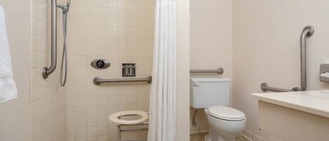 Un baño pequeño y accesible con inodoro, barras de apoyo, ducha con cabezal de ducha manual y lavamanos con espejo.