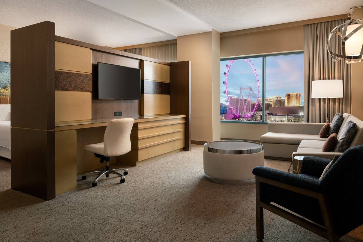 Habitación de hotel con escritorio, silla, sofá y un ventanal con vistas a una noria.