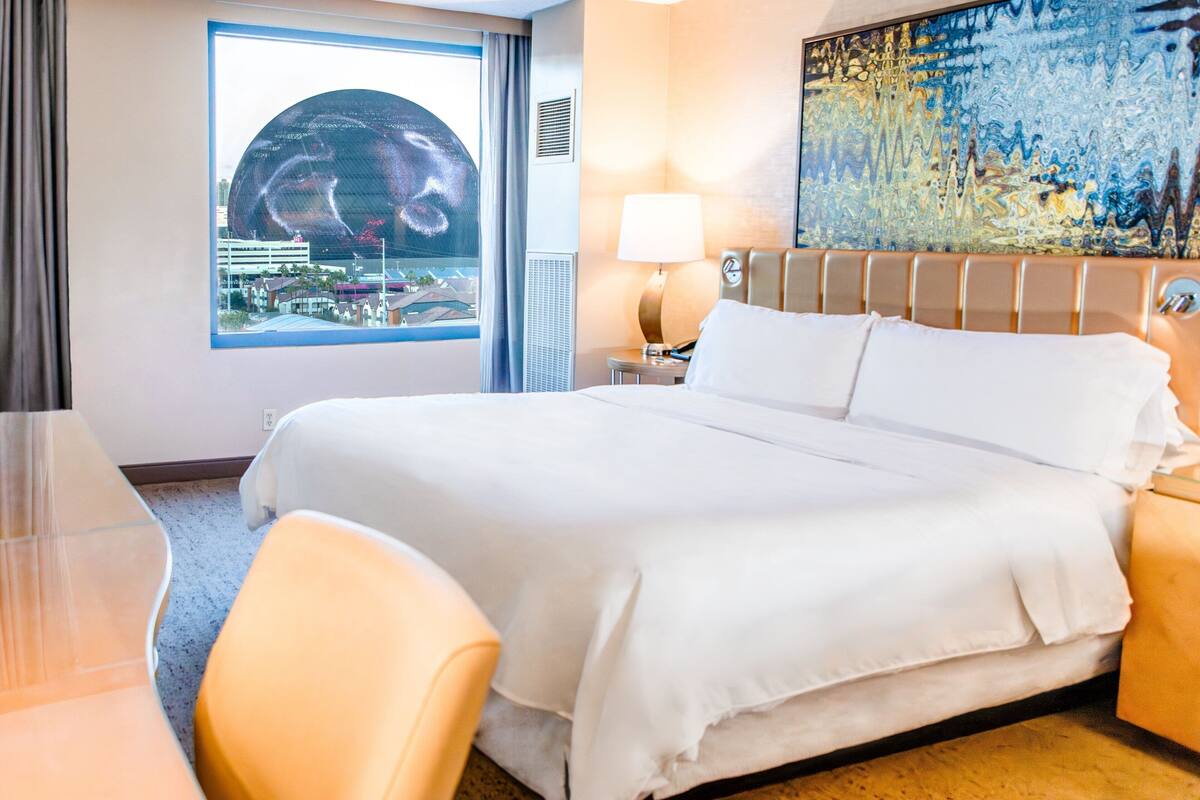 Habitación de hotel con una cama grande, un escritorio y una silla. Se ve una ventana con vistas a edificios y un cuadro en la pared.