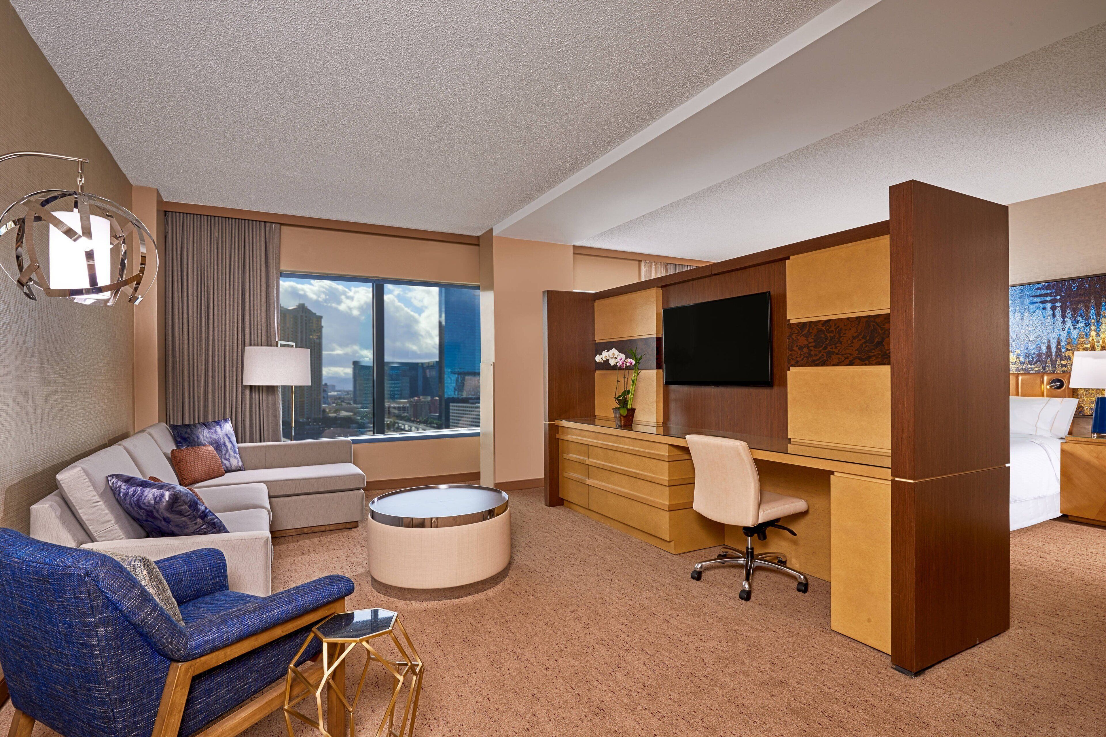 A modern hotel room with a sofa, round coffee table, desk, and TV.