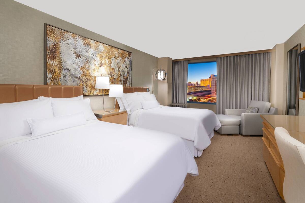 Habitación de hotel con dos camas, un escritorio, una silla y un ventanal con vistas a la ciudad.