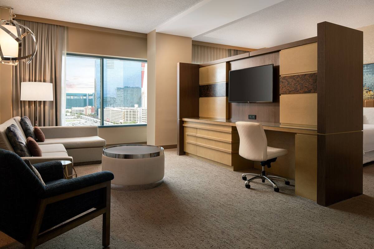 Una habitación de hotel moderna con un ventanal grande, un sofá, un escritorio, una silla y un televisor.