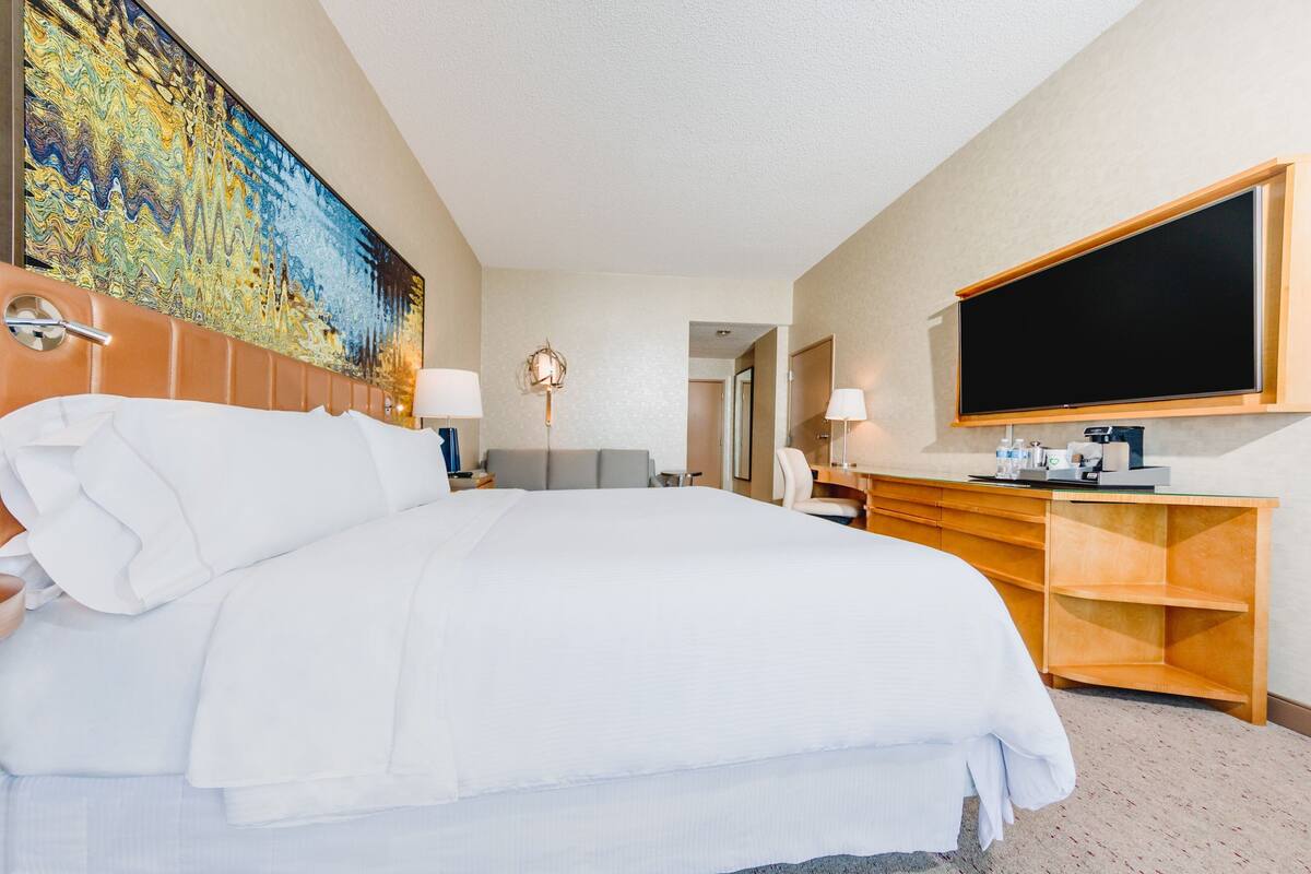Habitación de hotel con una cama grande, un escritorio con televisor de pantalla plana, un cuadro en la pared y un baño visible a través de una puerta abierta.