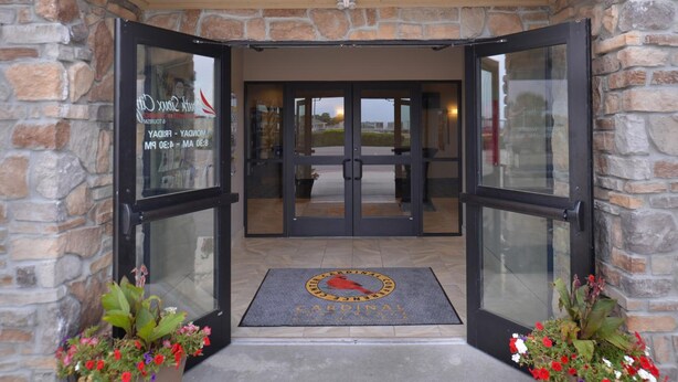 Entrada de hotel com paredes de pedra e portas de vidro, um tapete de boas-vindas e vasos de flores.