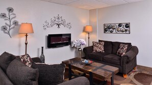 Uma sala de estar com sofá, poltronas, mesa de centro, duas luminárias, televisão de tela plana, arte decorativa na parede e um vaso com flores.