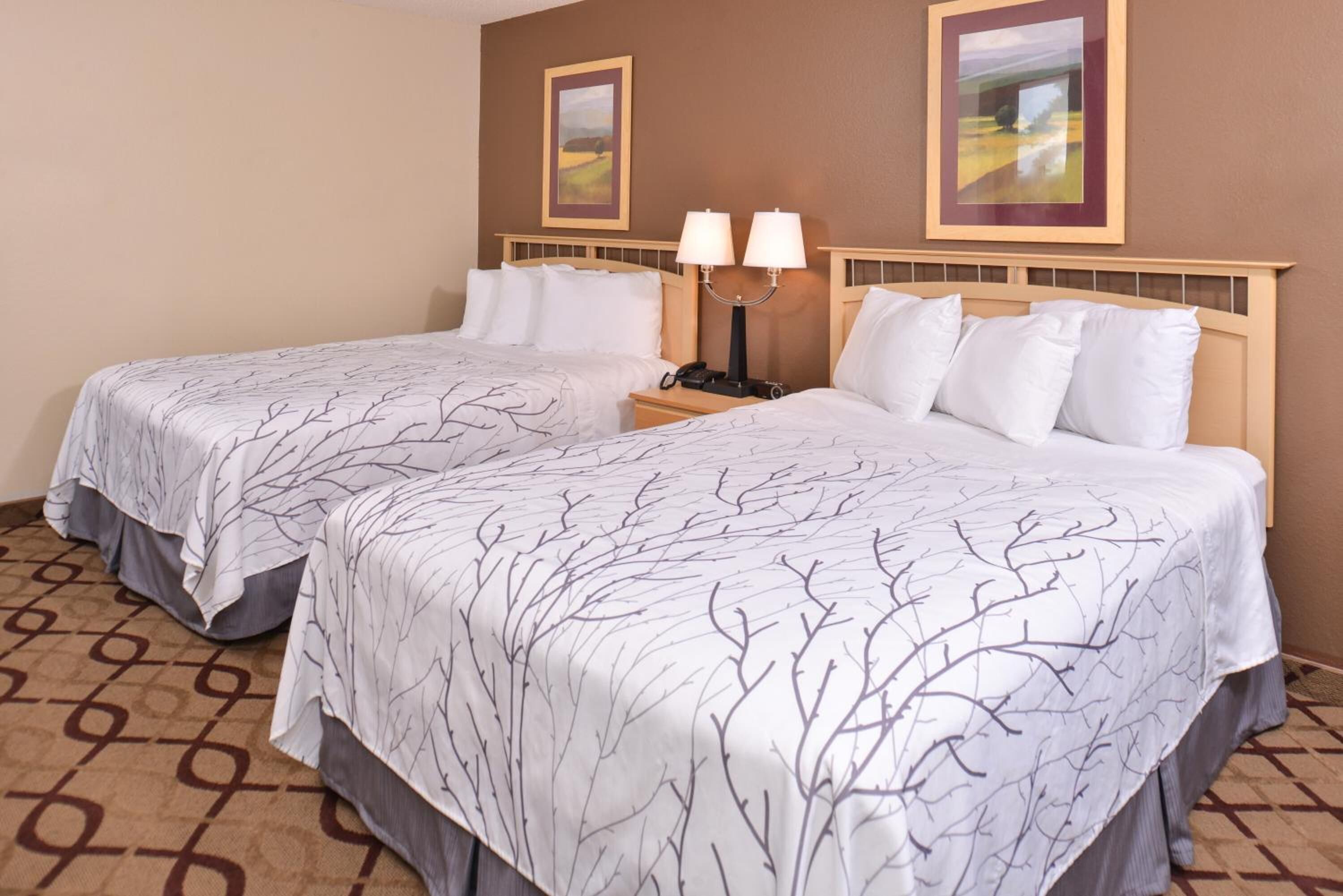 A hotel room with two beds, each with a tree branch pattern bedspread, a nightstand with a lamp, and two framed pictures on the wall.