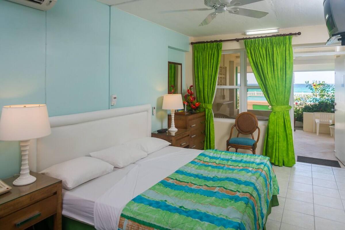 Habitación de hotel con una cama, una mesita de noche con lámpara, un aparador, una silla y vista al océano a través de una ventana con cortinas verdes.