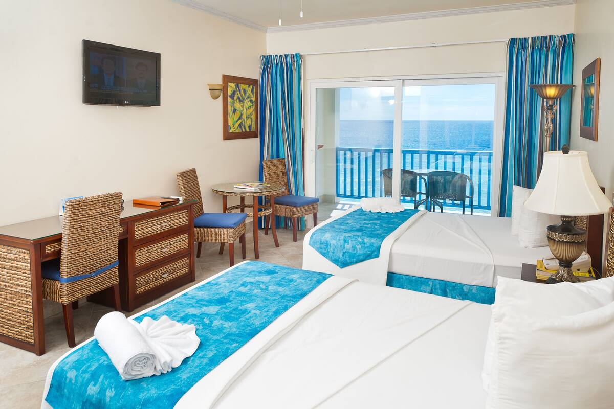 Habitación de hotel con dos camas, un escritorio, sillas, televisión y balcón con vista al mar.