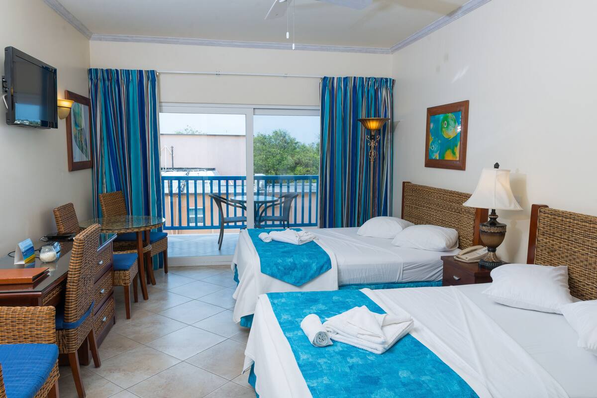 Habitación de hotel con dos camas, una mesa de comedor con sillas, un televisor y un balcón con vistas.