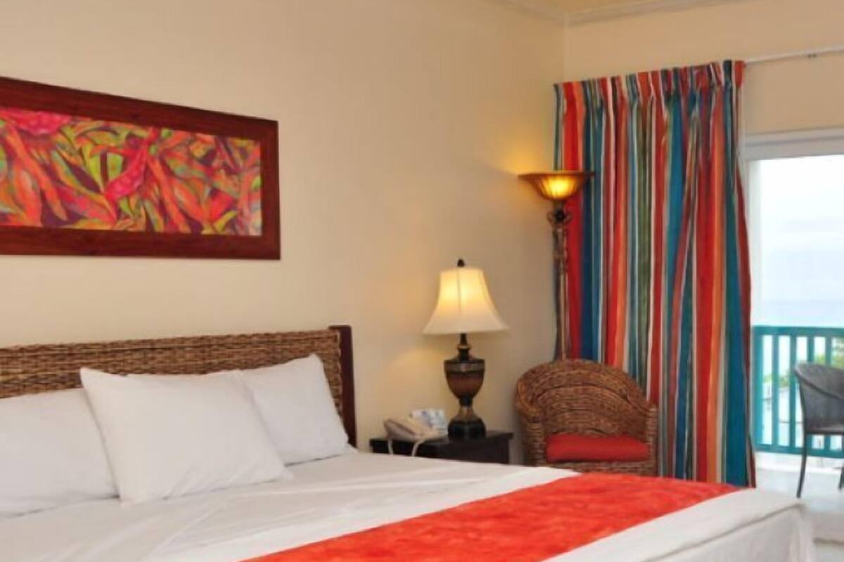 Una habitación de hotel con cama, mesita de noche, lámpara, silla, cortina colorida y un cuadro en la pared.
