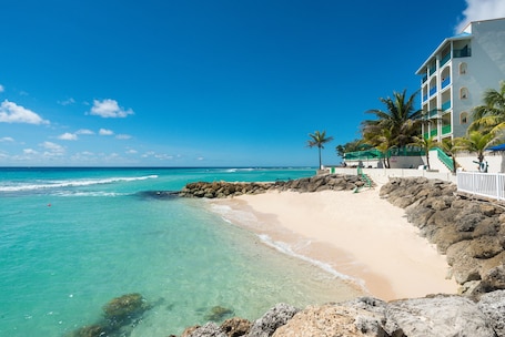 A beach with clear turquoise water, a sandy shore, and a building with balconies and a green railing.