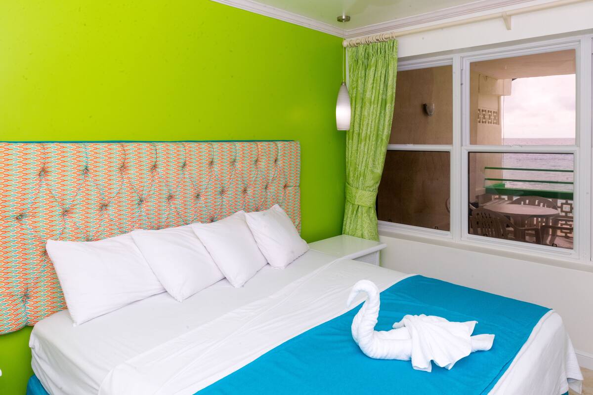 Habitación de hotel con pared verde brillante, cabecera estampada, ropa de cama blanca, un detalle azul en la cama y vista al mar a través de una ventana.