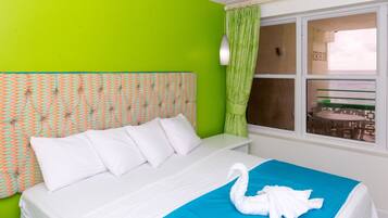 Habitación de hotel con pared verde brillante, cabecera estampada, ropa de cama blanca, un detalle azul en la cama y vista al mar a través de una ventana.