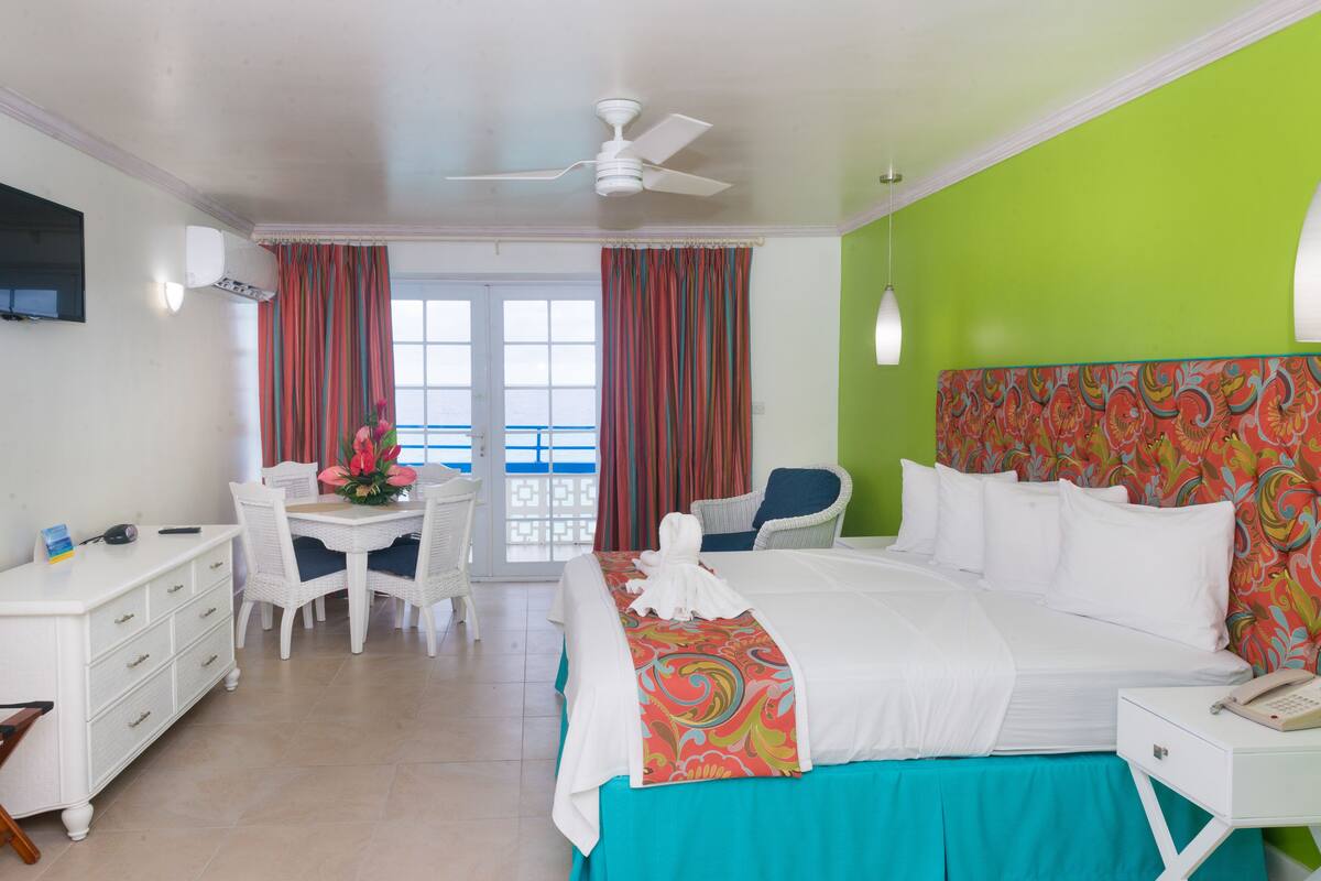 Habitación de hotel con cama, escritorio, silla, mesa con flores, balcón con cortinas y pared verde.