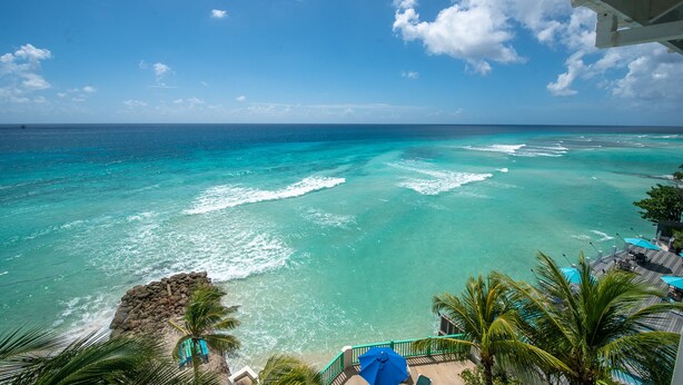 Vista costera con aguas turquesas cristalinas, una costa rocosa y una zona de resort con sombrillas y palmeras.