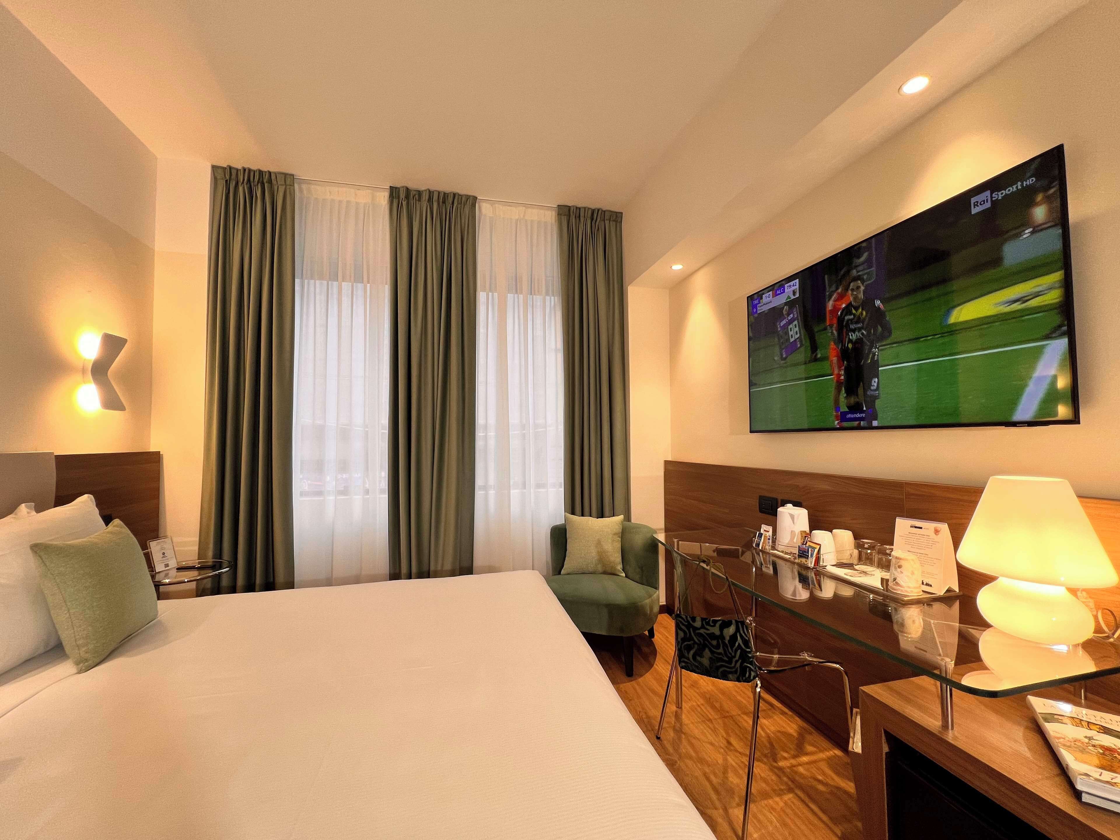 A hotel room with a large bed, a TV mounted on the wall, a desk with a lamp, and a chair.