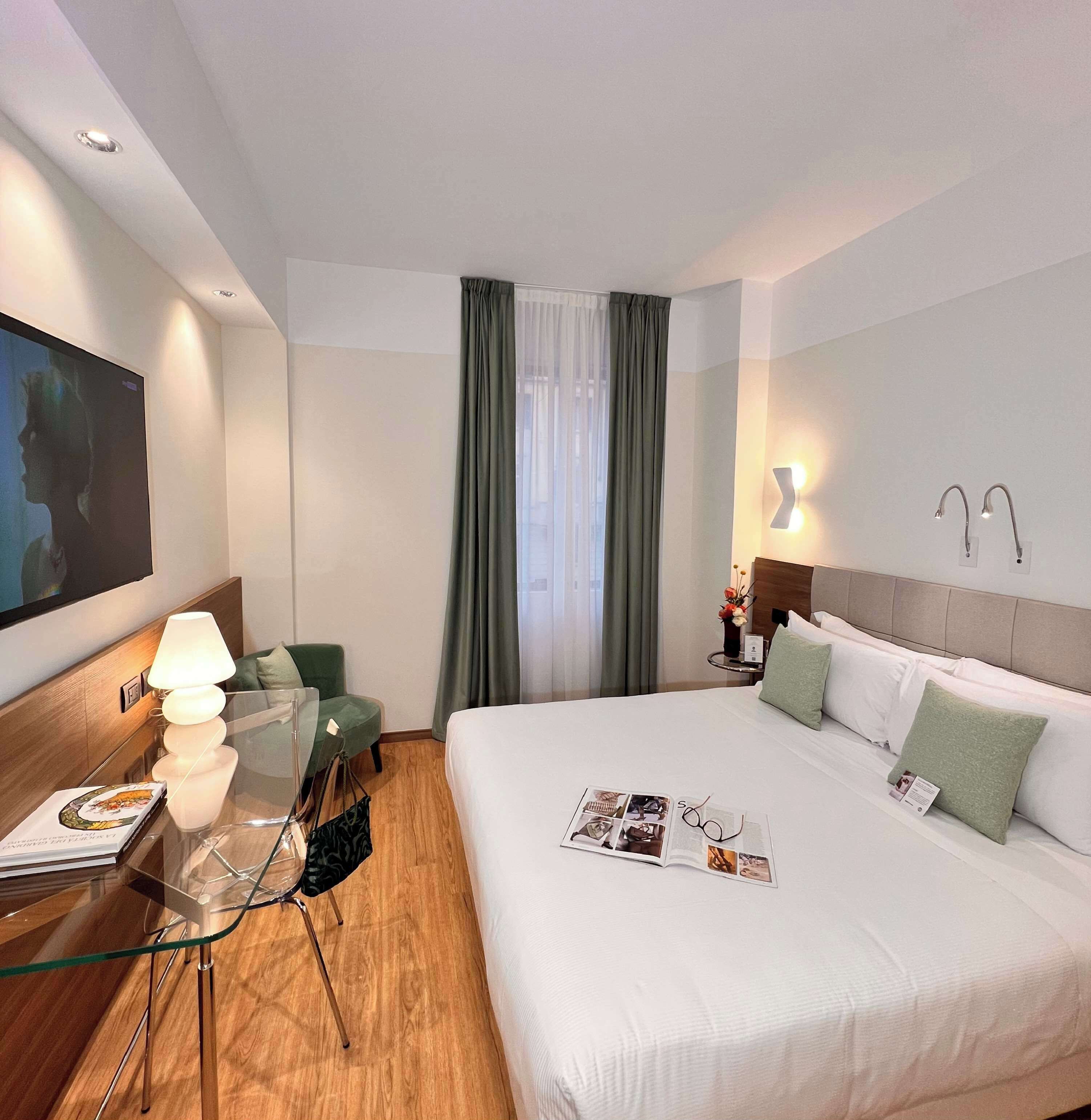 Ein Hotelzimmer mit Bett, Schreibtisch mit Lampe, Stuhl, Fernseher und einem Fenster mit Vorhängen.