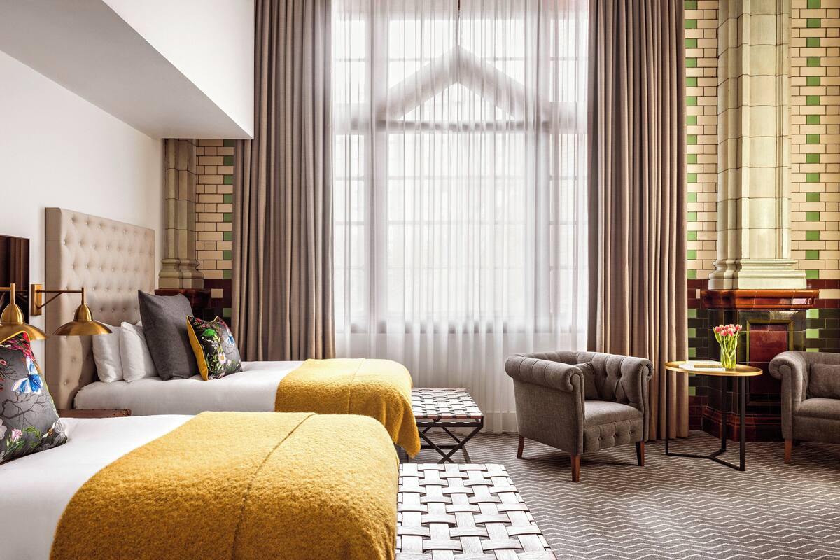 A hotel room with two beds, a sofa, a chair, a small table, and a large window with curtains.