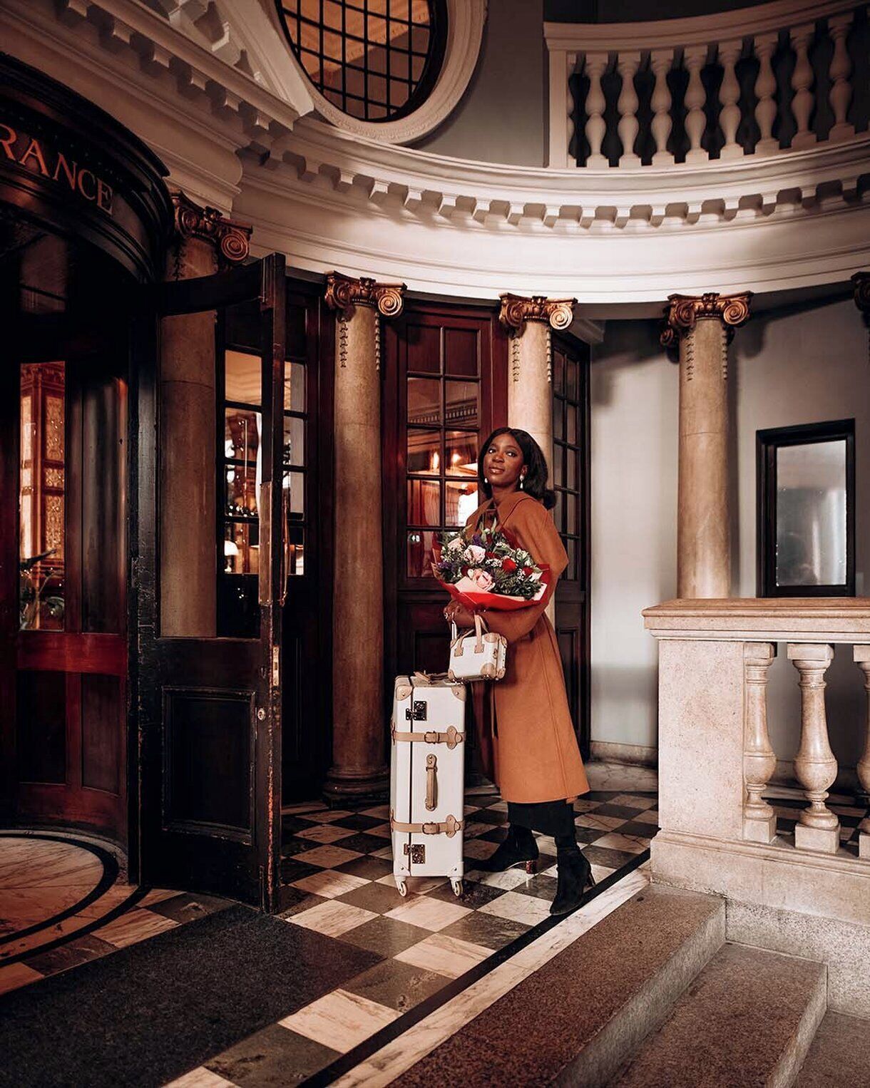 A grand entrance with a checkered floor, a woman holding a bouquet, and a vintage suitcase.