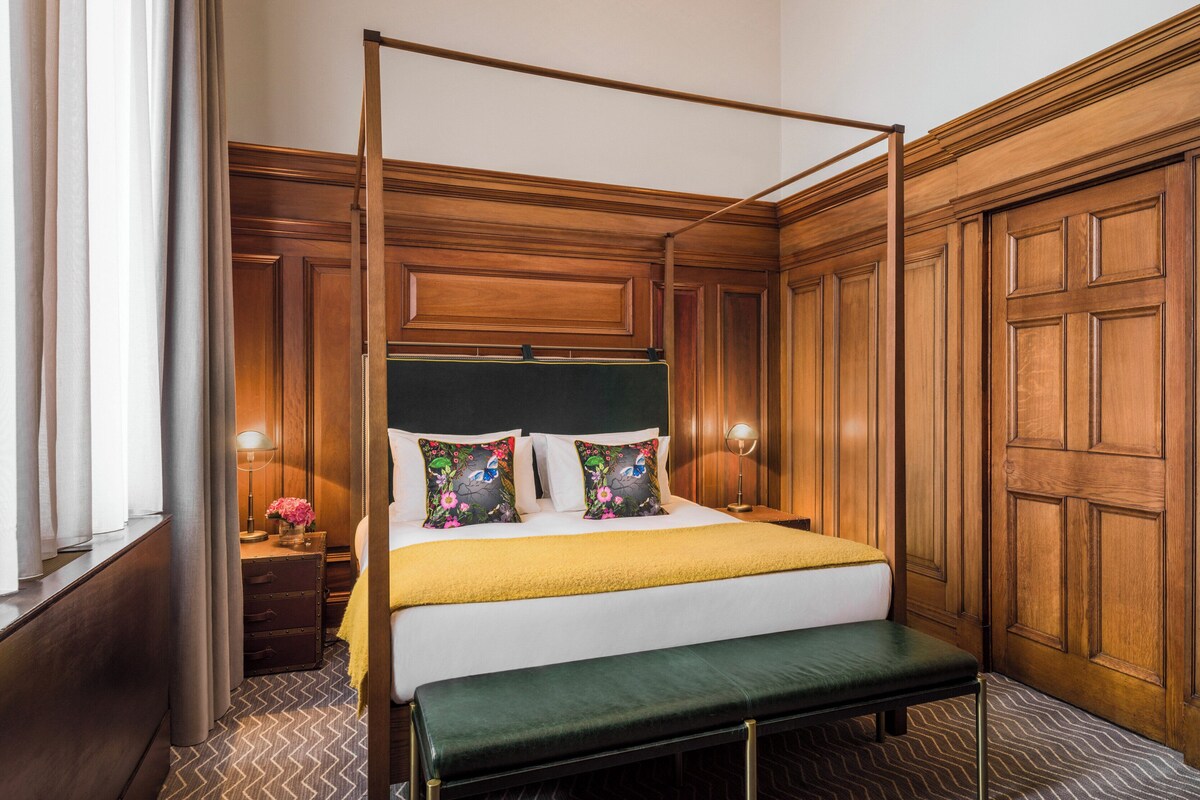 A four-poster bed with a green upholstered headboard, a bench at the foot of the bed, and a bedside table with a lamp and flowers.