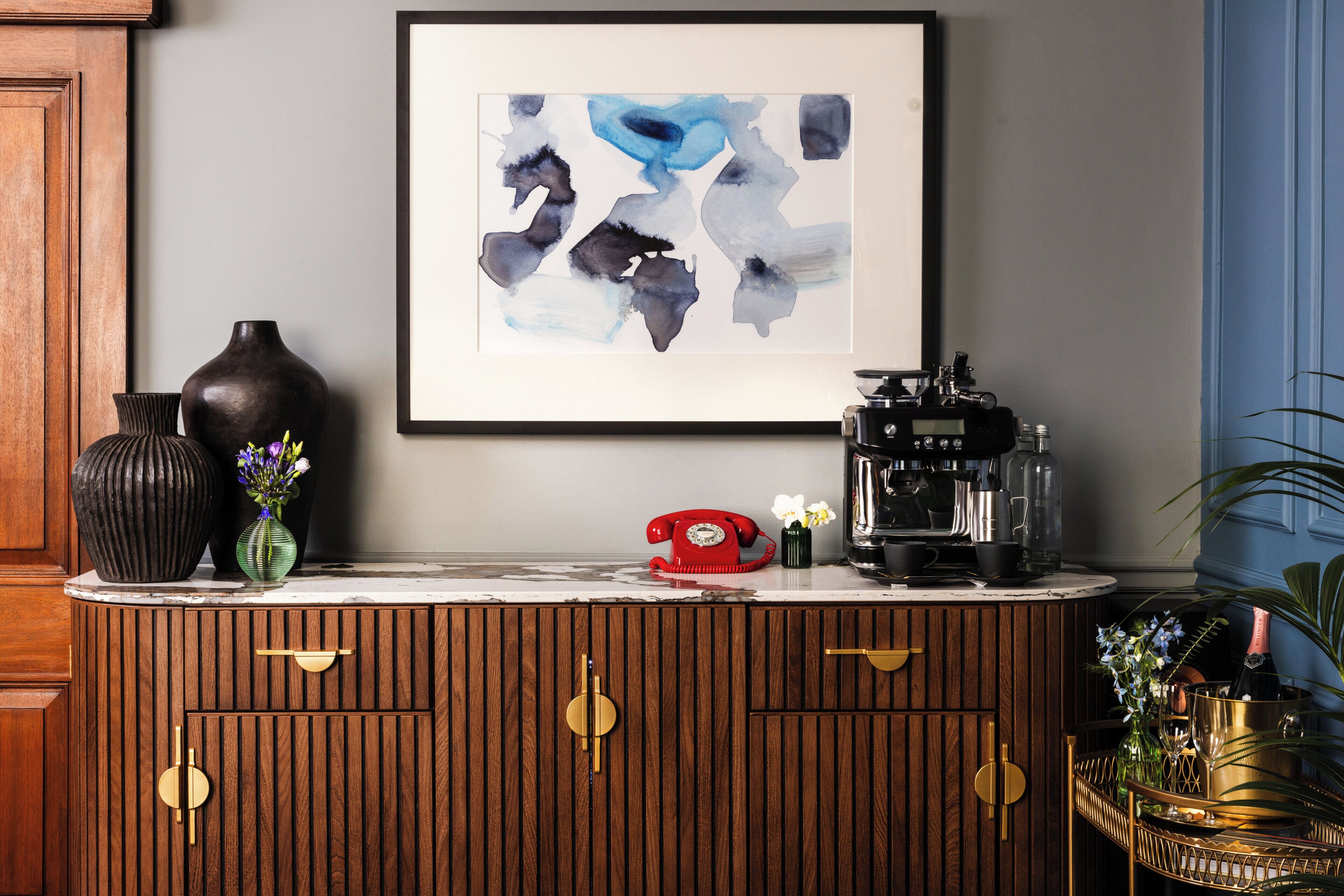 A wooden cabinet with drawers, a framed abstract painting, a vintage red telephone, a coffee machine, and decorative vases.