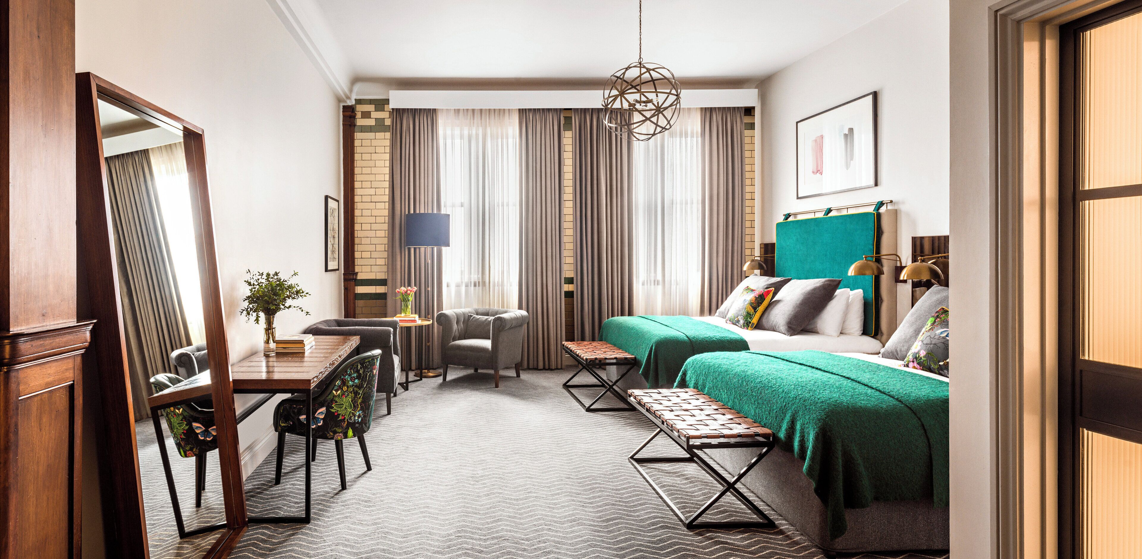 A hotel room with a bed, a desk, a chair, a table with a vase, and a hanging light fixture.