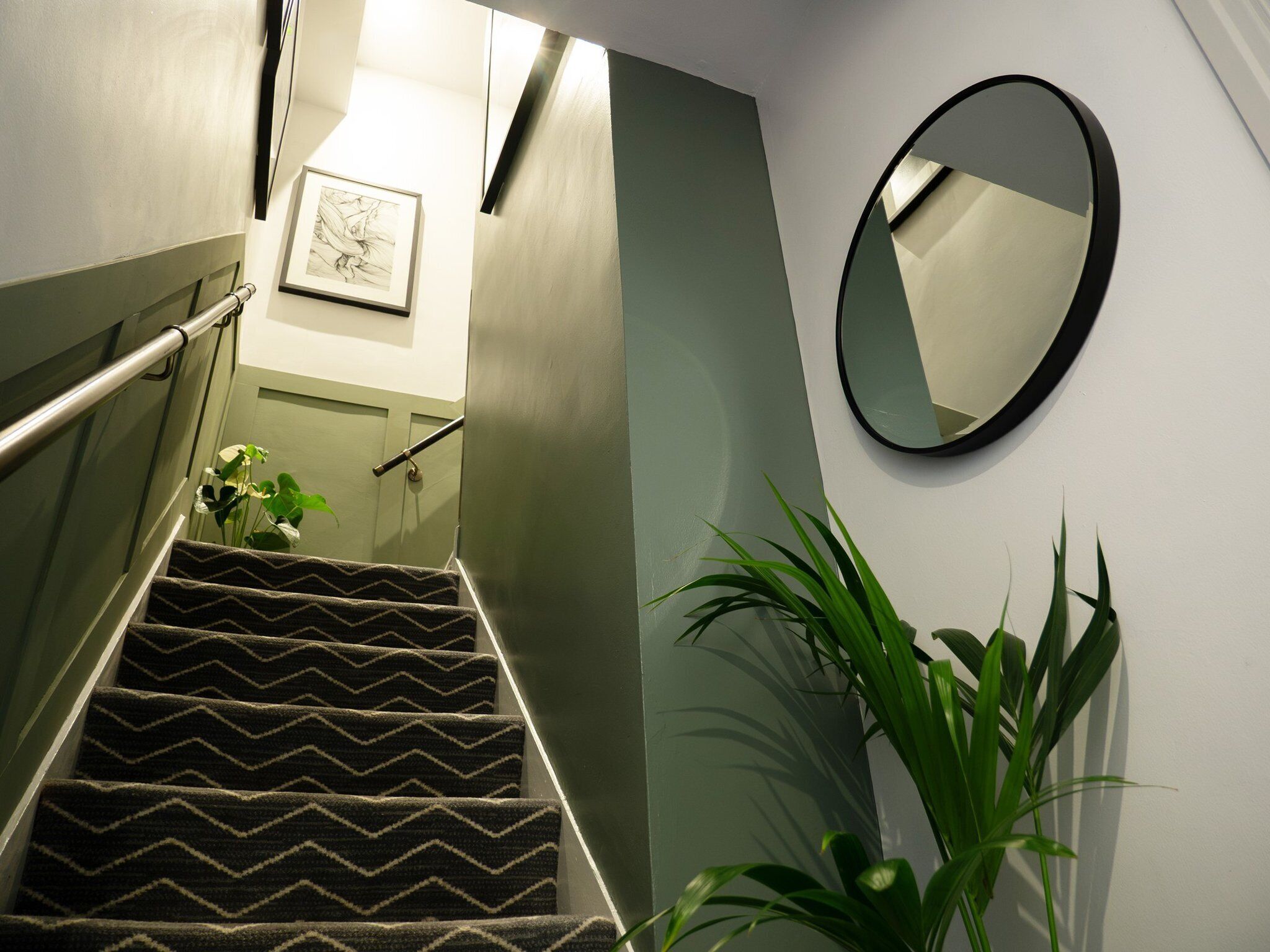 A staircase with a zigzag carpet pattern, a metal handrail, a framed artwork on the wall, a potted plant, and a round mirror.