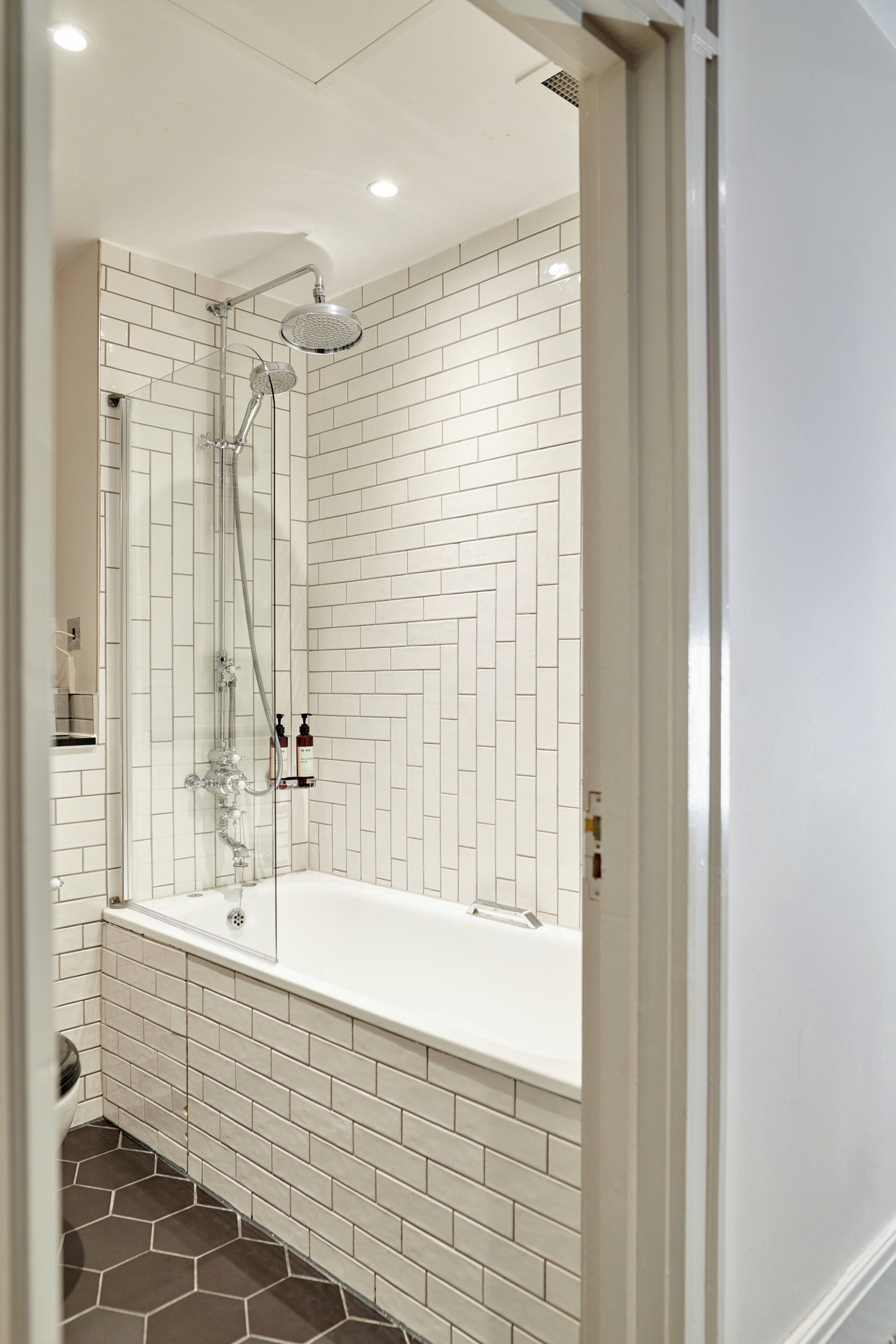 Ein modernes Badezimmer mit weißen, zickzack-verlegten Fliesen, einer gläsernen Duschkabine und einer weißen Badewanne.