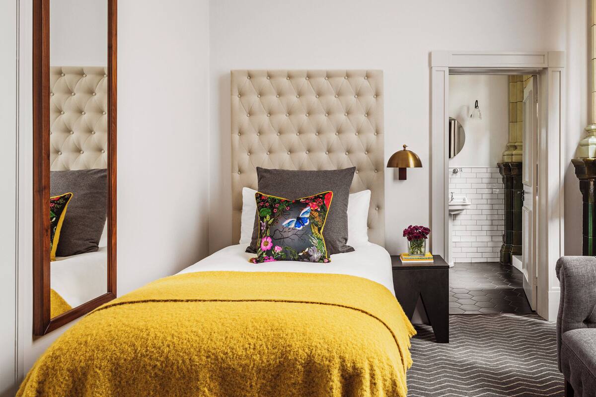 A hotel room with a tufted headboard, a bed with a mustard-colored blanket, a grey armchair, and a view of the bathroom through an open door.