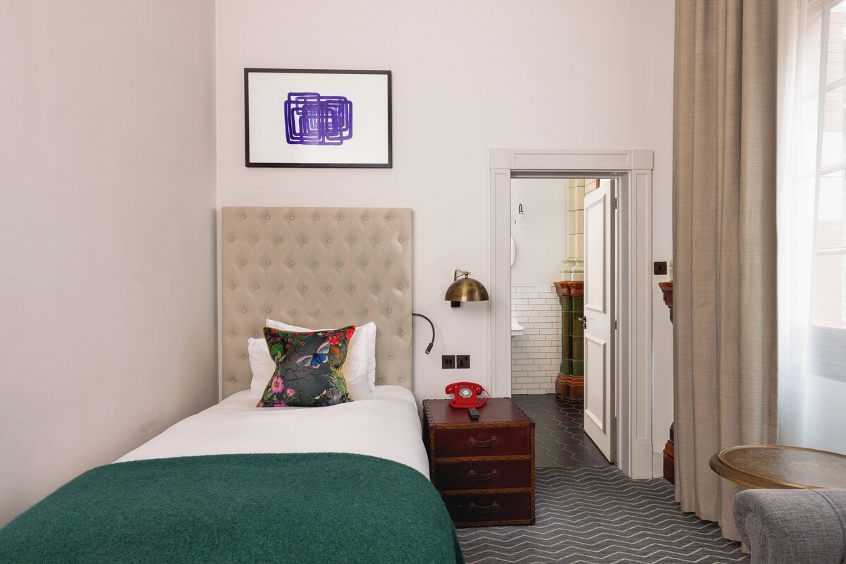 Habitació d'hotel amb un llit, una taula de nit amb un telèfon, una làmpada penjada a la paret i una fotografia emmarcada sobre el llit.
