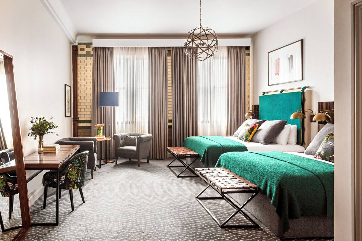A hotel room with a bed, a desk with a chair, a small table with a vase, and a bench.