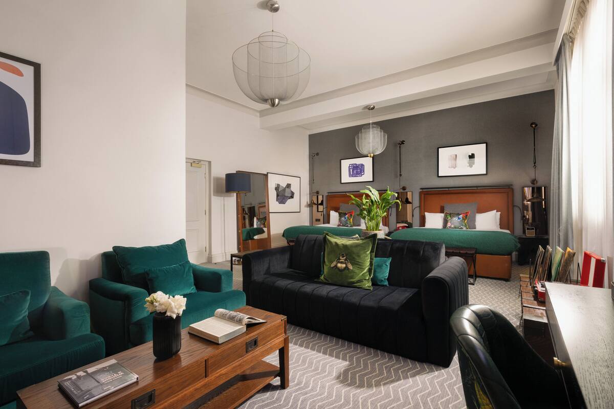 A modern hotel room with a green sofa, a wooden coffee table, a bed with a green headboard, and a large window with curtains.
