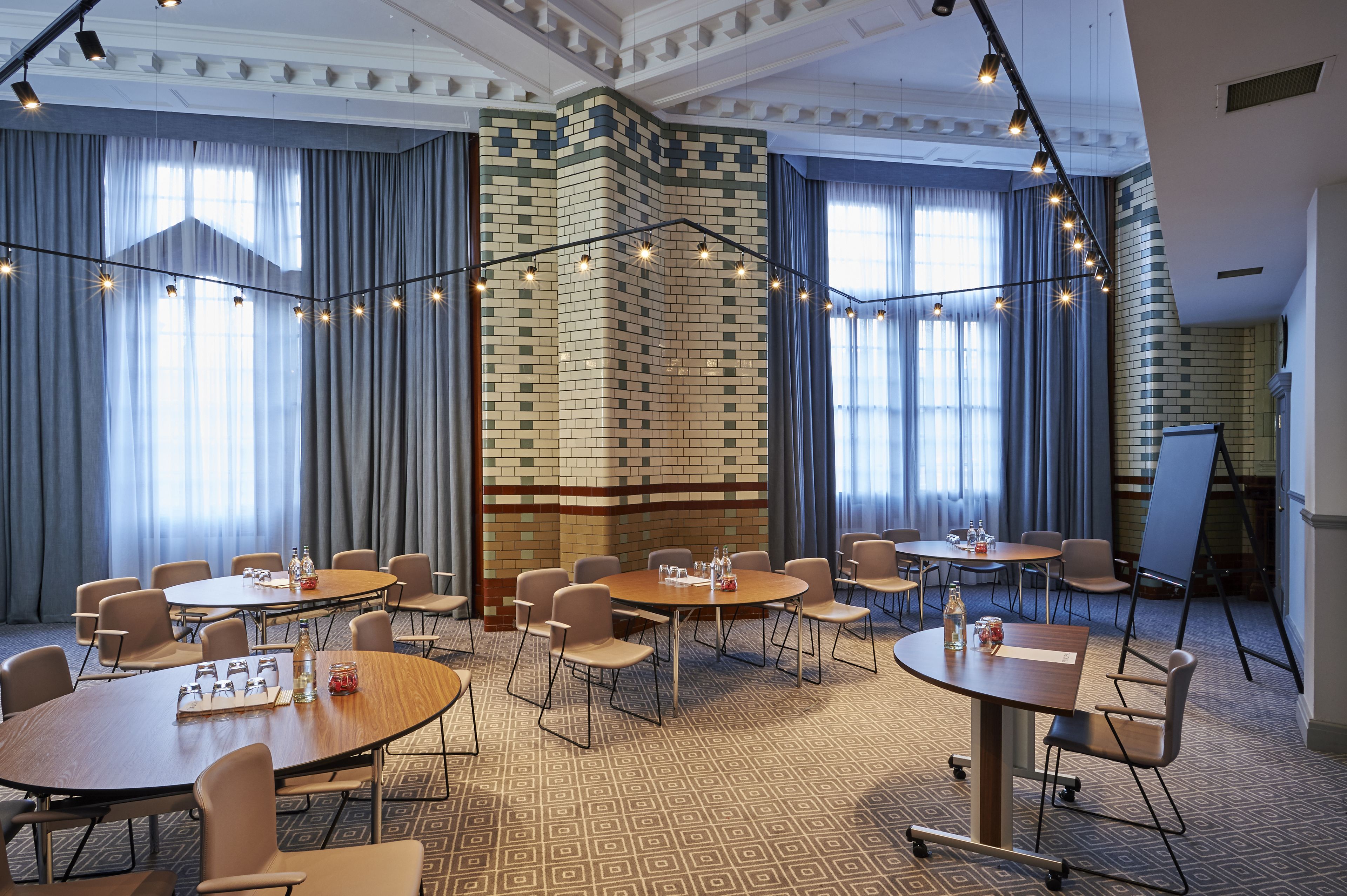Une salle de conférence dotée de plusieurs tables et chaises, d’un mur décoratif en briques et de grandes fenêtres avec des rideaux.