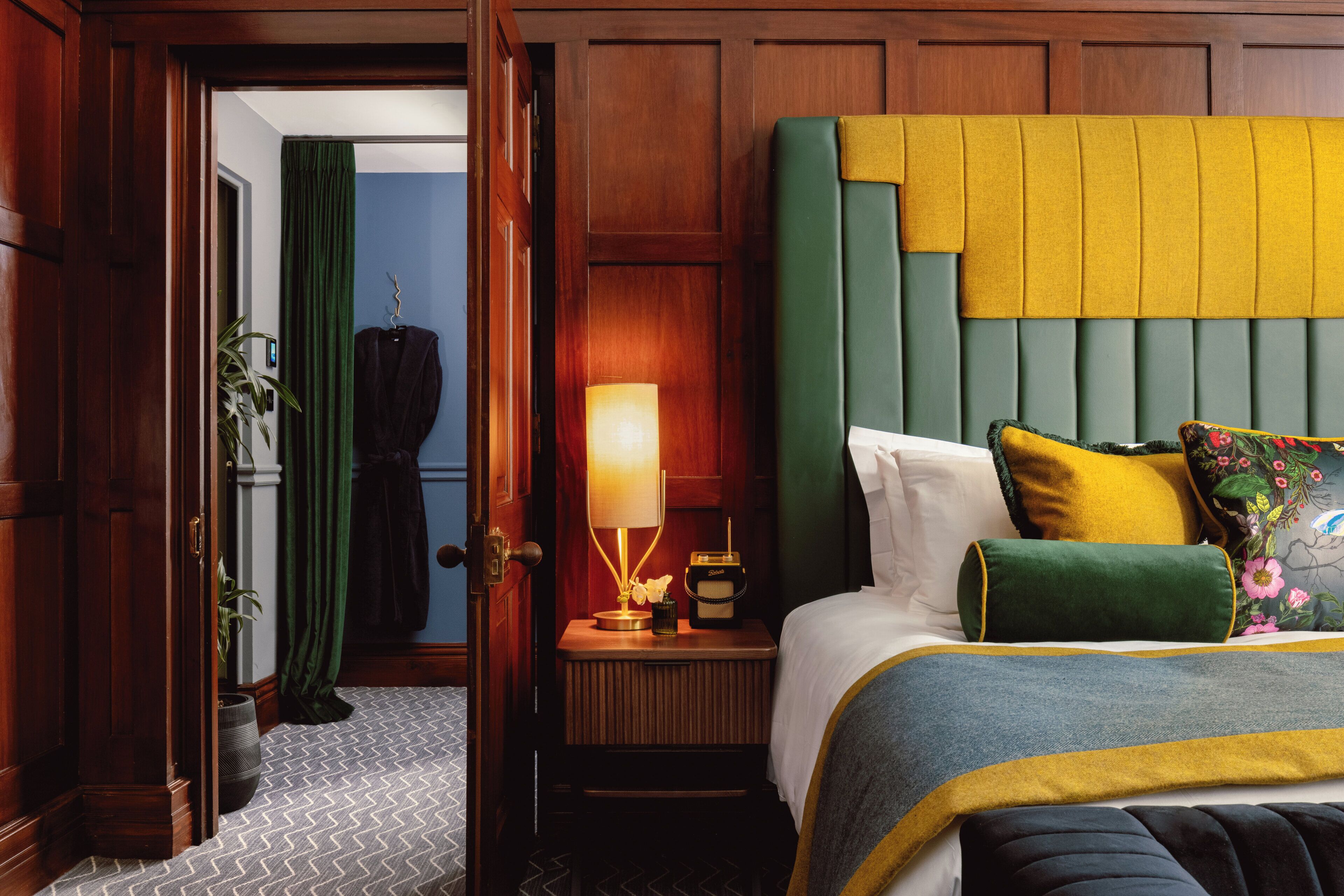 A hotel room with a large bed, a nightstand with a lamp, a potted plant, and a wooden door.