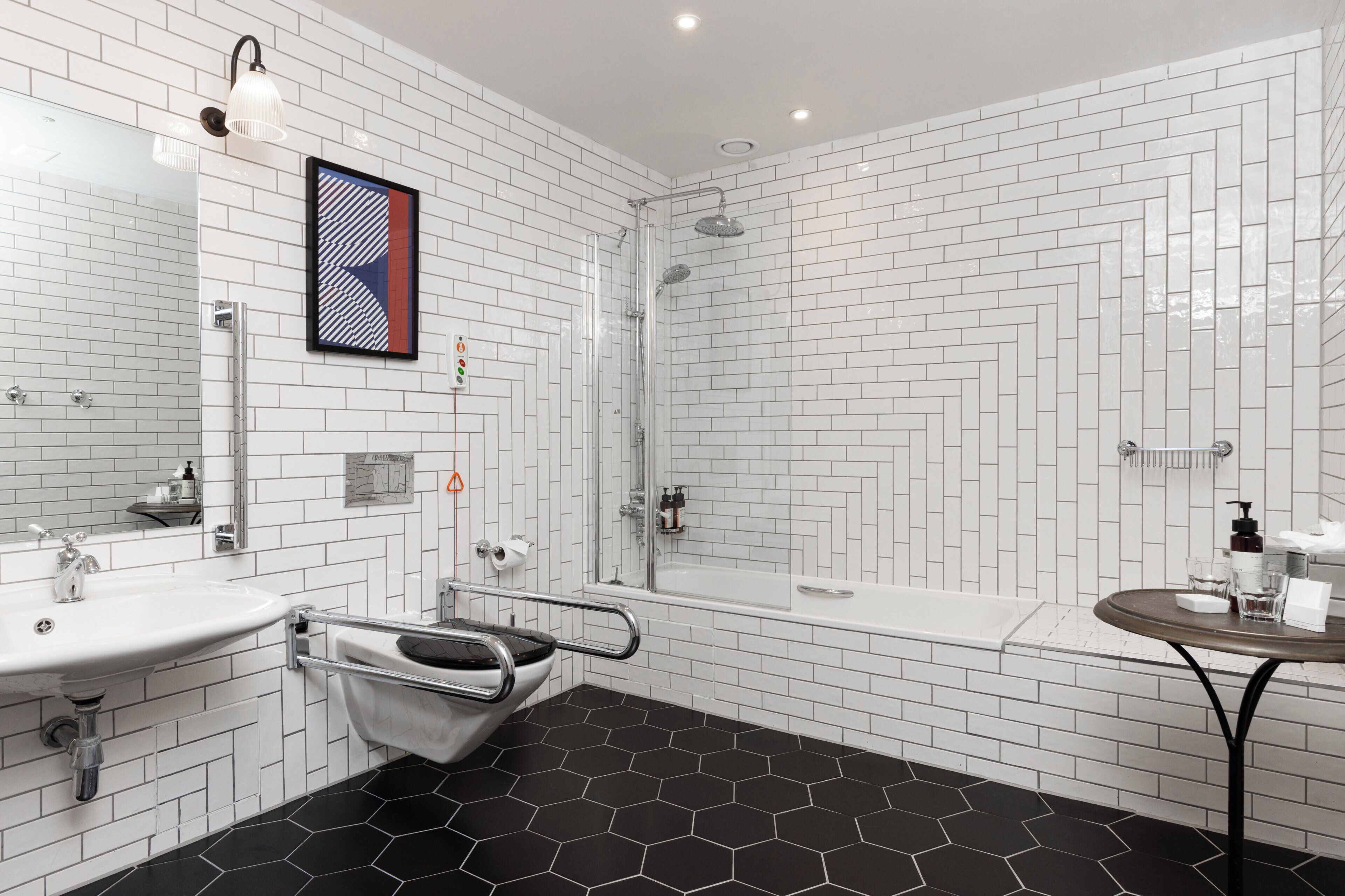 Un baño moderno con azulejos blancos tipo metro, piso hexagonal negro, ducha, lavamanos e inodoro con barra de apoyo.