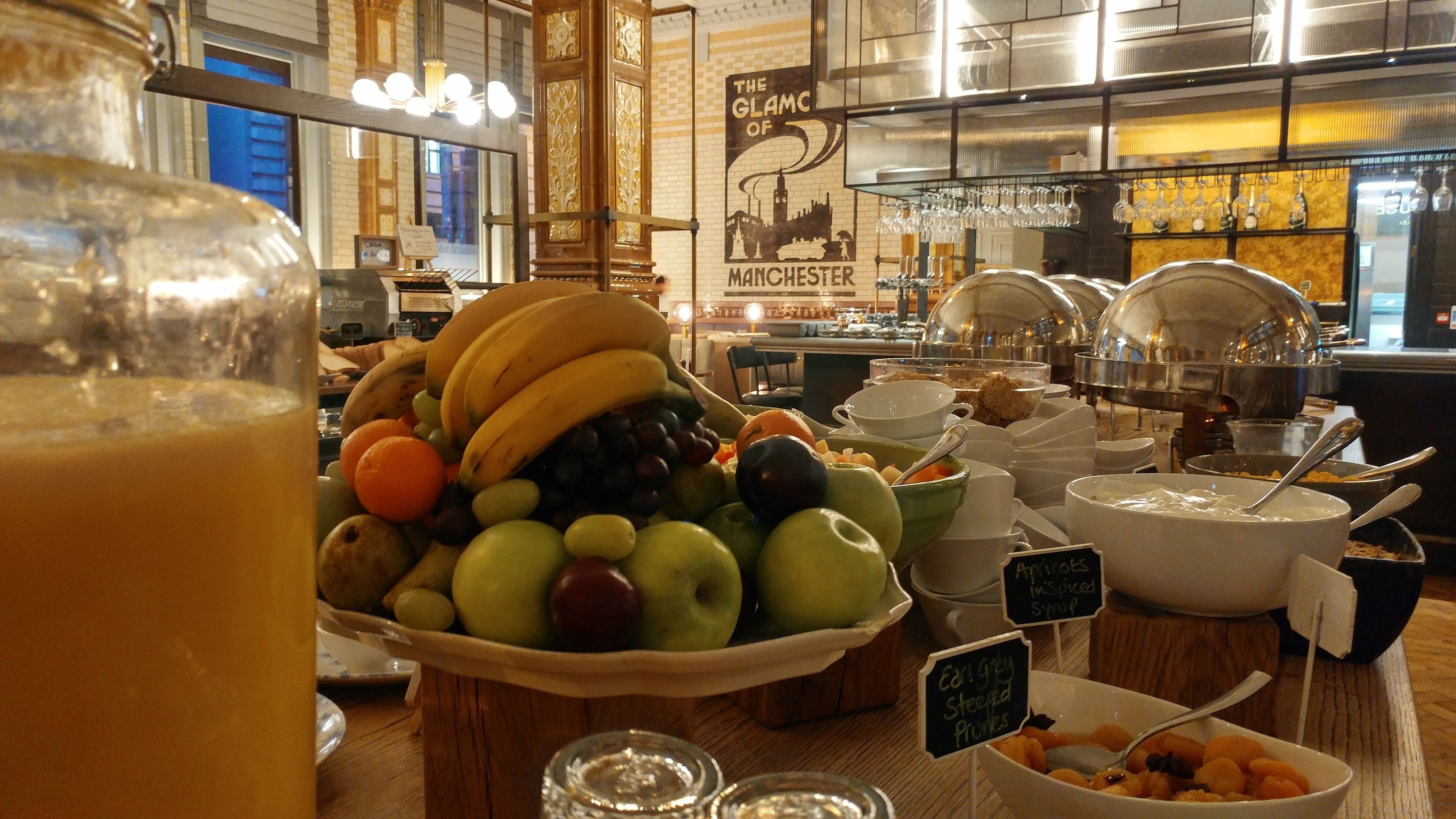 A buffet with various fruits and bowls of food, including apples and apricots, in a well-lit dining area with a mural of Manchester.